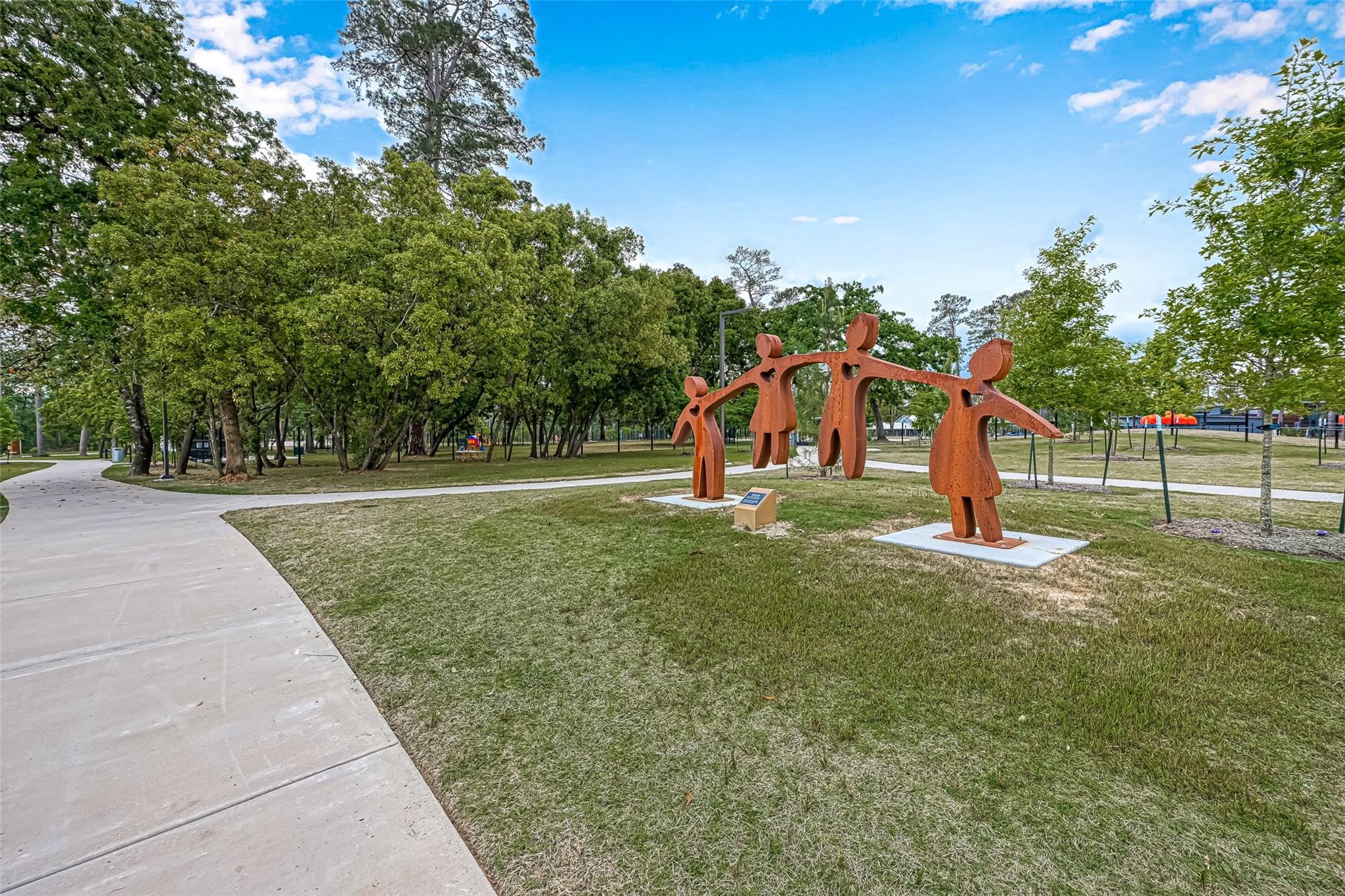 Neighborhood park with a curving path, mature trees, and a rust-colored sculpture of four linked figures.