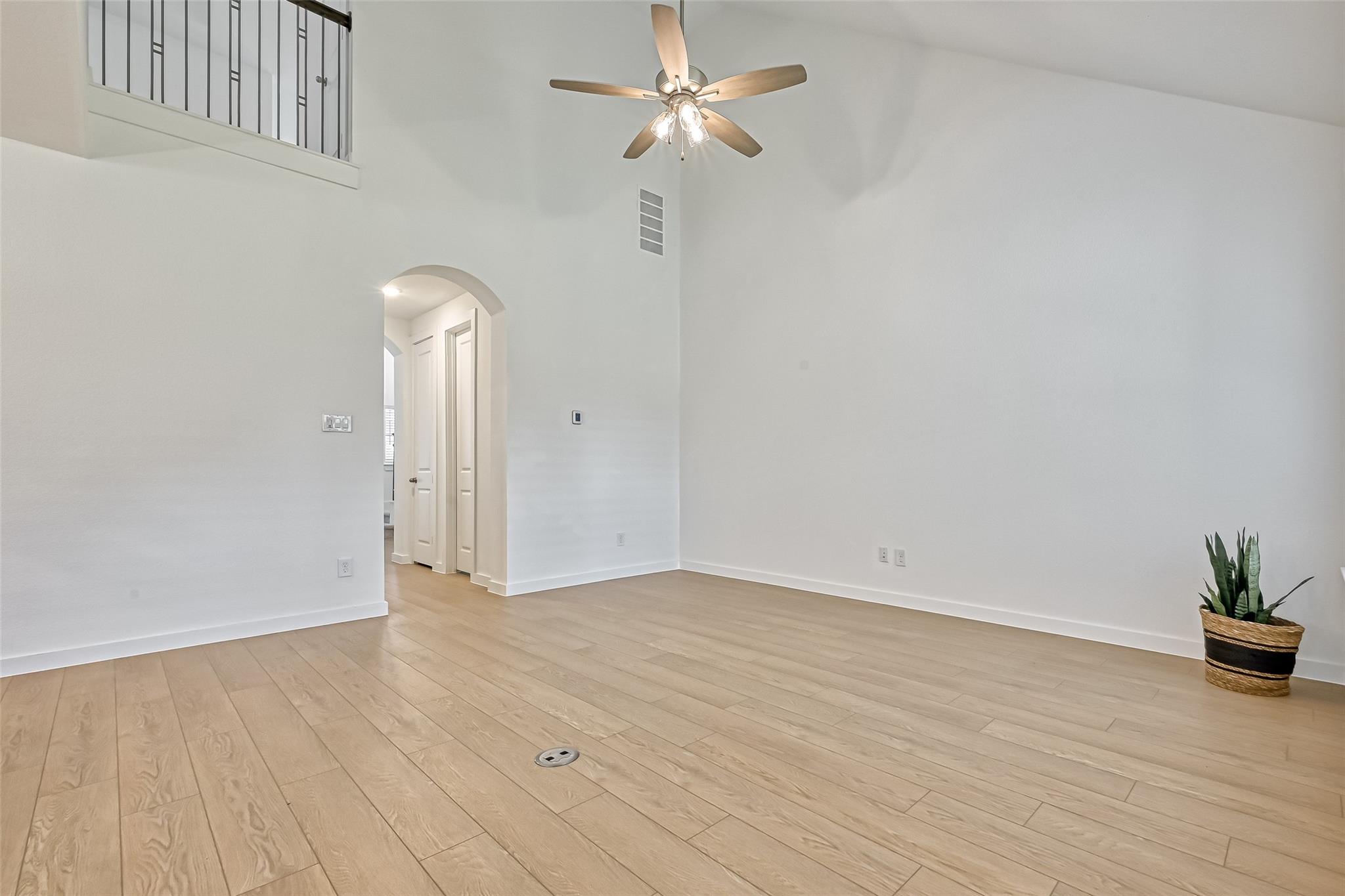 Mostly empty living room with high ceilings, light plank flooring, a ceiling fan, and an upstairs railing.