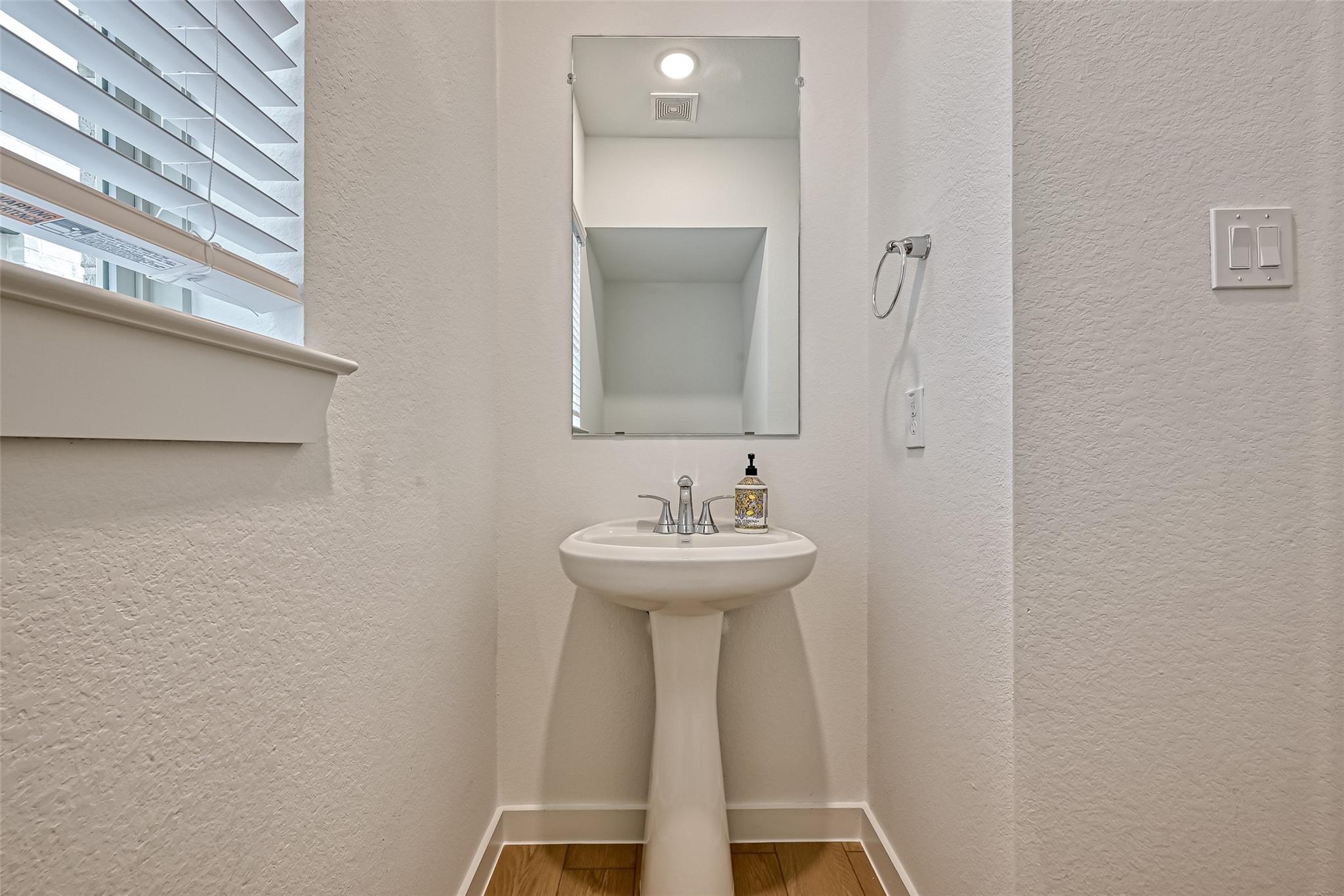 Powder room with a white pedestal sink, rectangular mirror, and window with horizontal blinds.