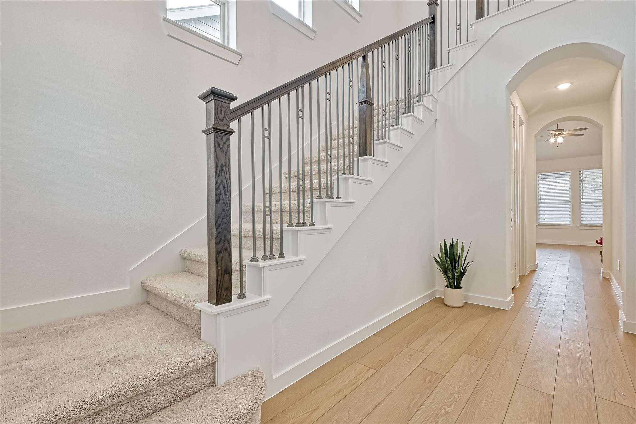 Entryway with carpeted staircase, dark railing, light wood floors, and an arched hallway.