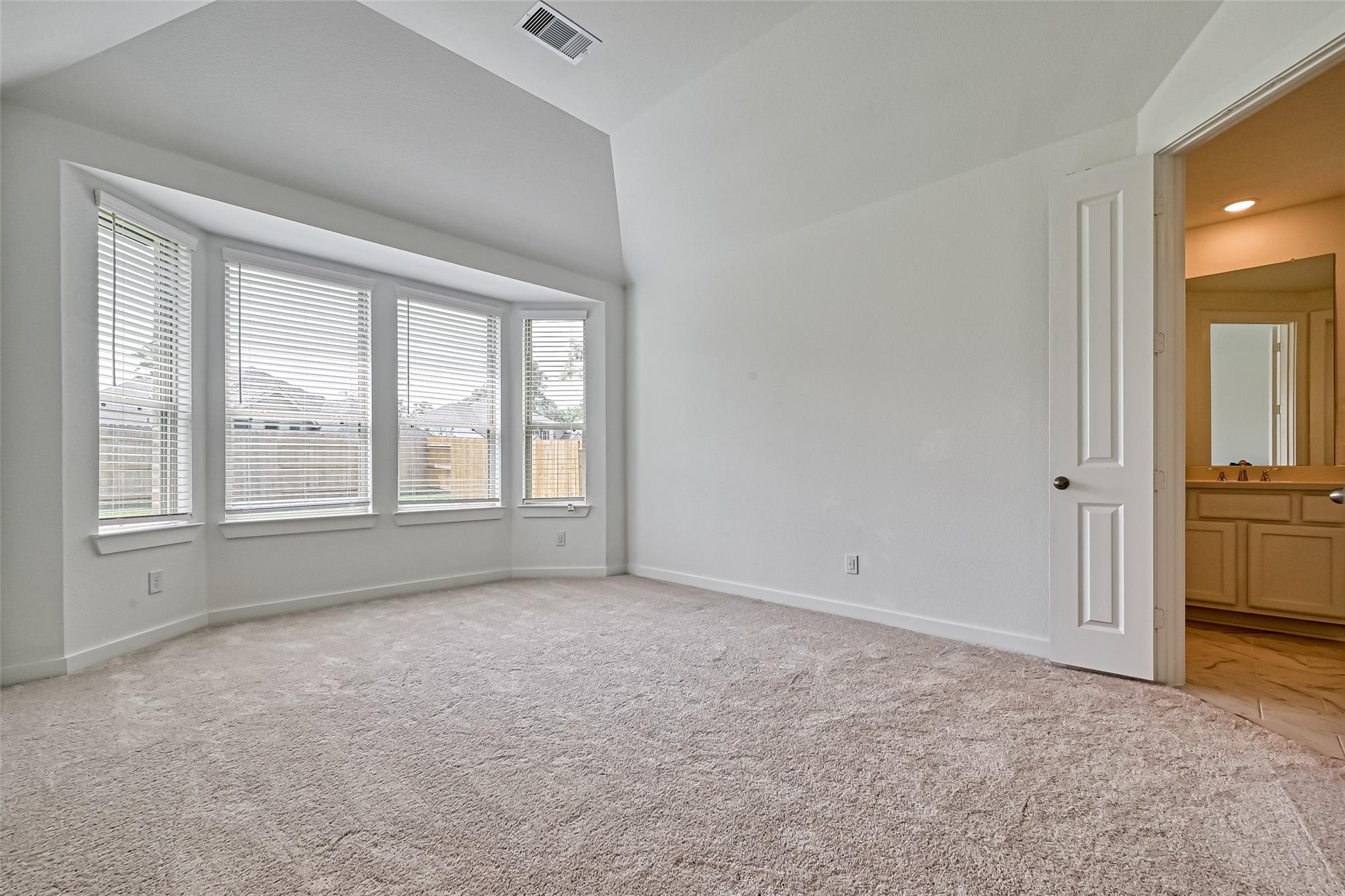 Empty carpeted bedroom with a vaulted ceiling, four windows, and an open doorway to the bathroom.