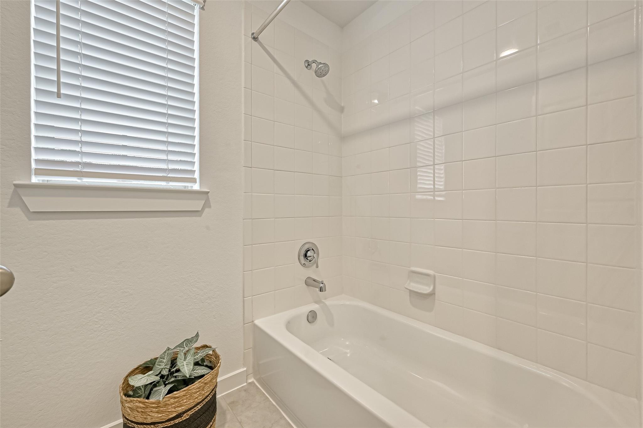 Bathroom bathtub with white tiled walls, chrome shower fixtures, window blinds, and a potted plant.