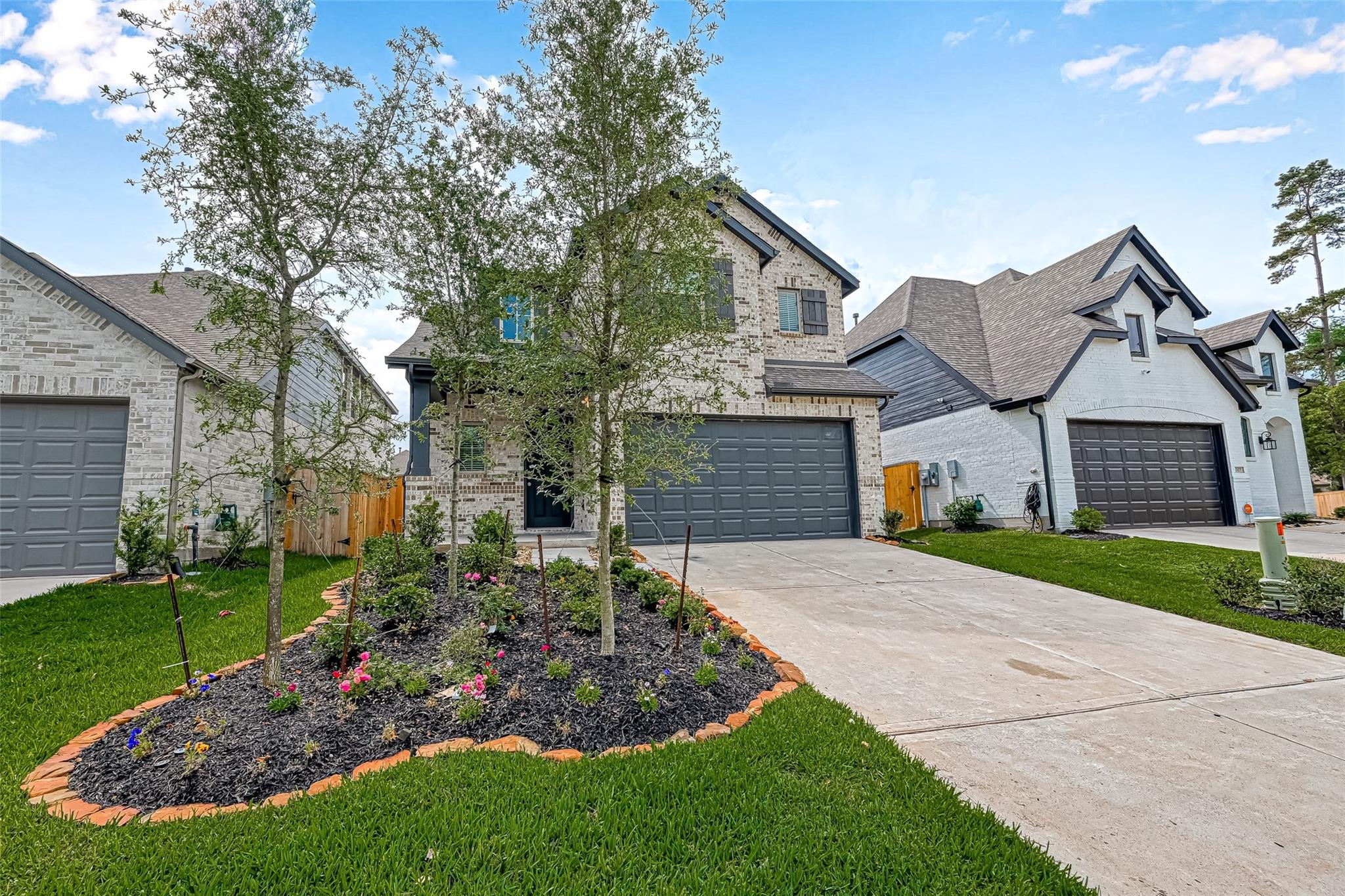 Two-story brick home with a wide gray garage door, concrete driveway, and landscaped garden with young trees and flowers.