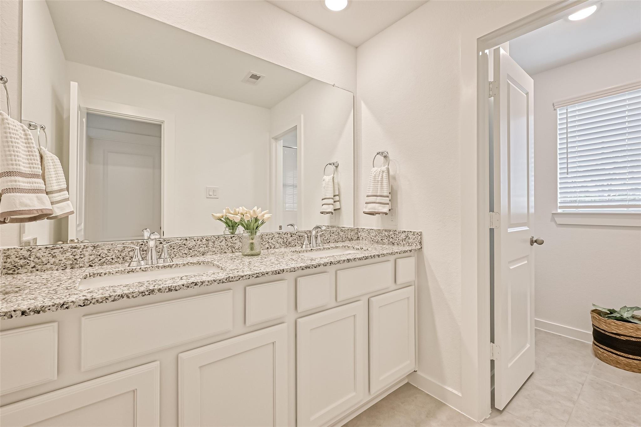 Bright bathroom with a double vanity, speckled granite counter, large mirror, and open door to a window.