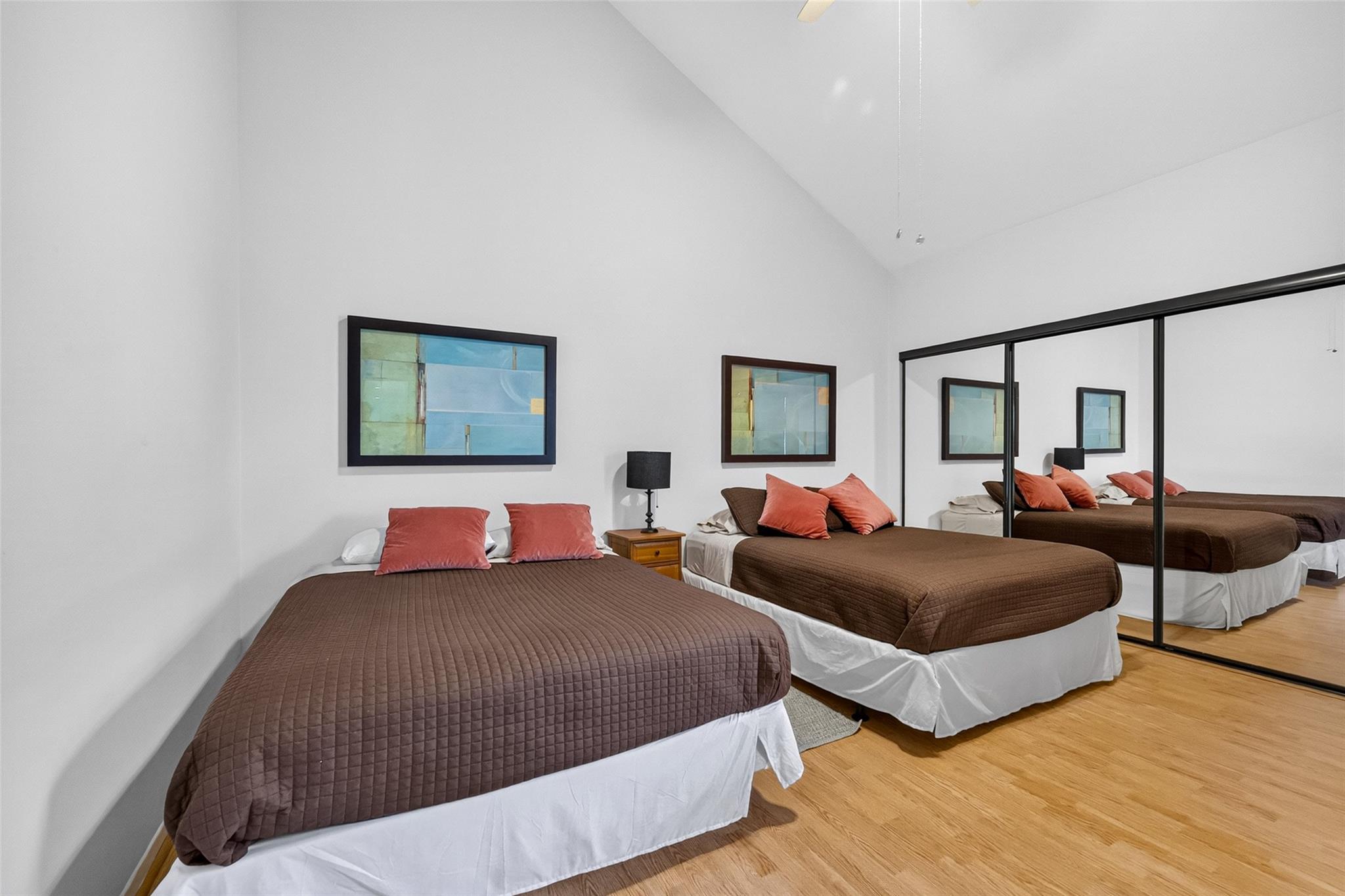 Bedroom with two brown-covered beds, coral pillows, mirrored closet doors, and hardwood floors.