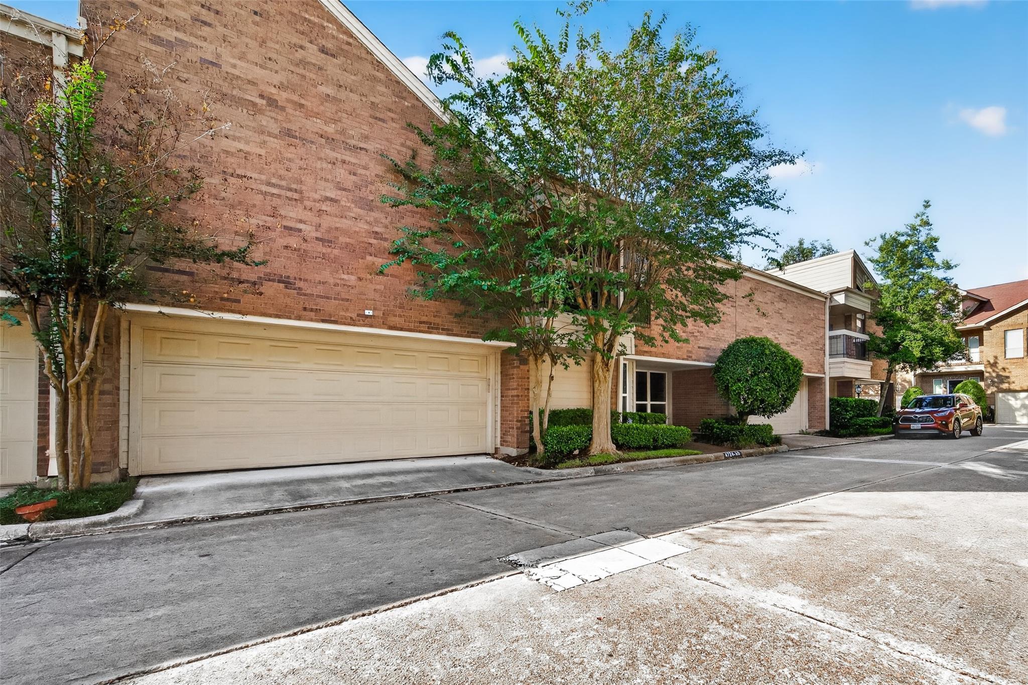 Brick condominium exterior with a large garage door, leafy trees, and a paved driveway.