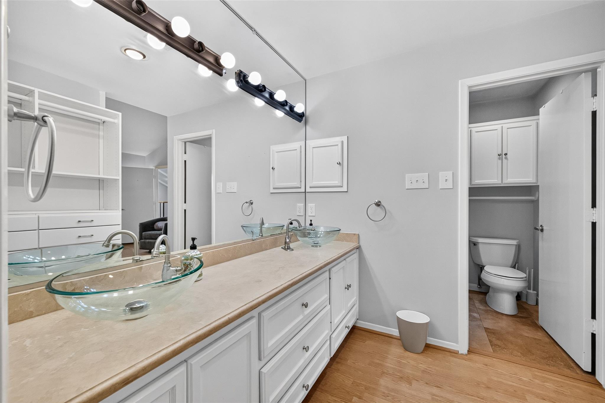 Bathroom with two glass vessel sinks, white cabinetry, a large mirror, and a separate toilet alcove.