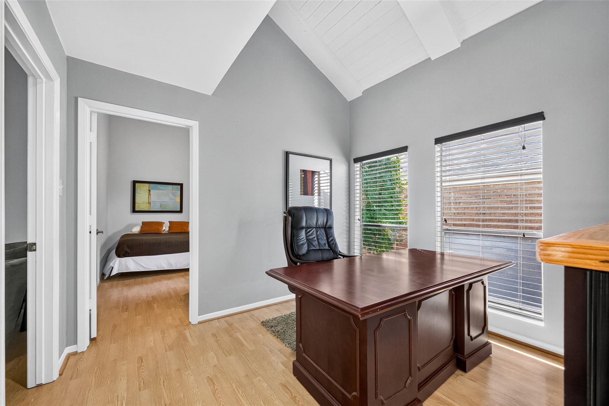 Furnished home office with a large wood desk, vaulted ceiling, and doorway to a bedroom.
