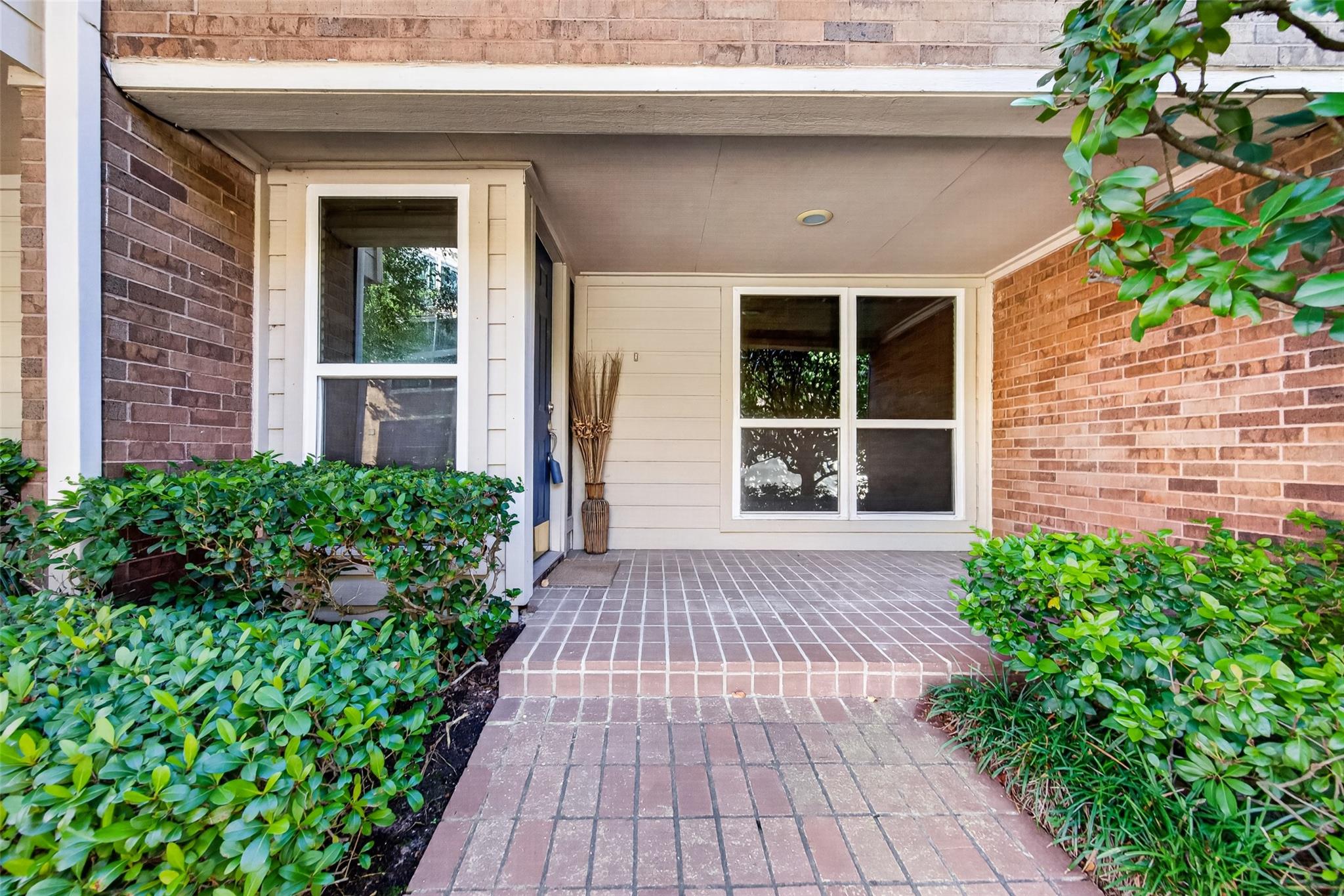 Covered brick entry porch with a paved walkway, white-framed windows, and shrubs.