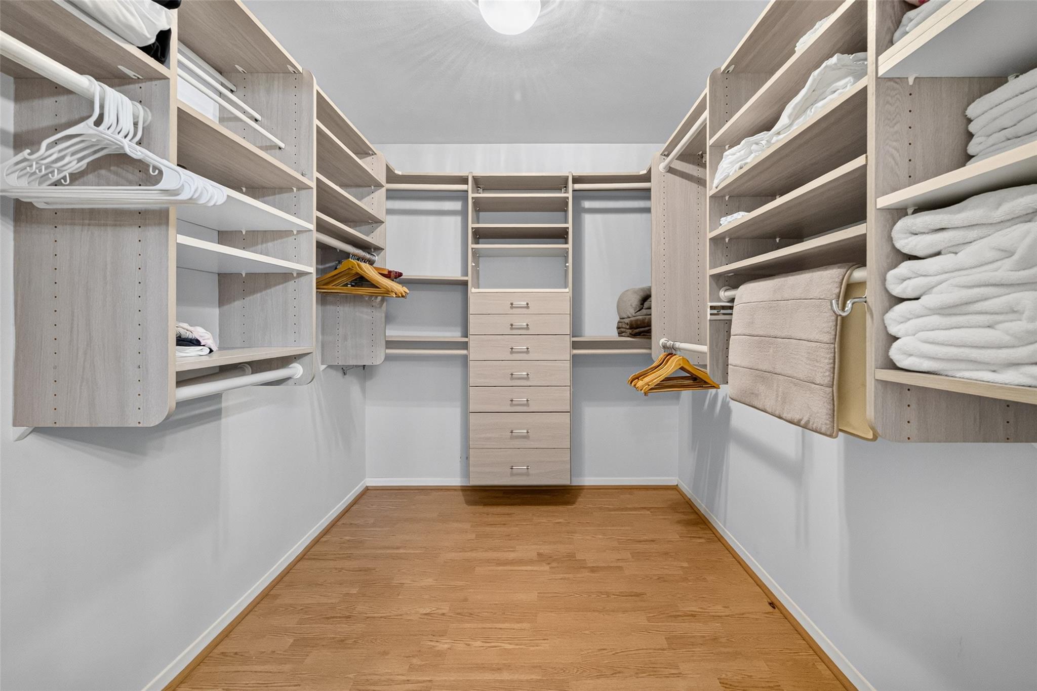 Walk-in closet with light wood shelving, central drawers, folded towels, and hardwood flooring.