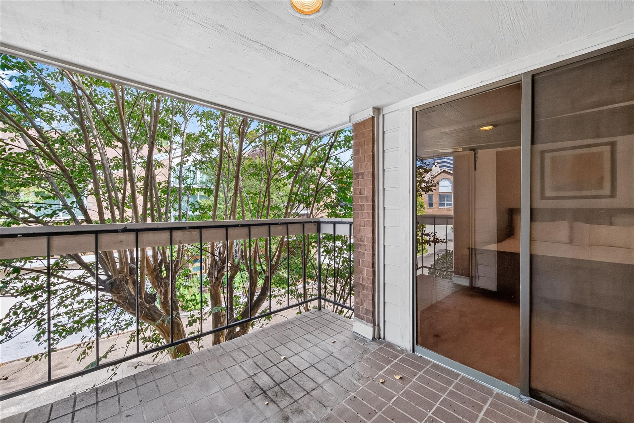 Covered condo balcony with tiled floor, black railing, and leafy trees beyond.