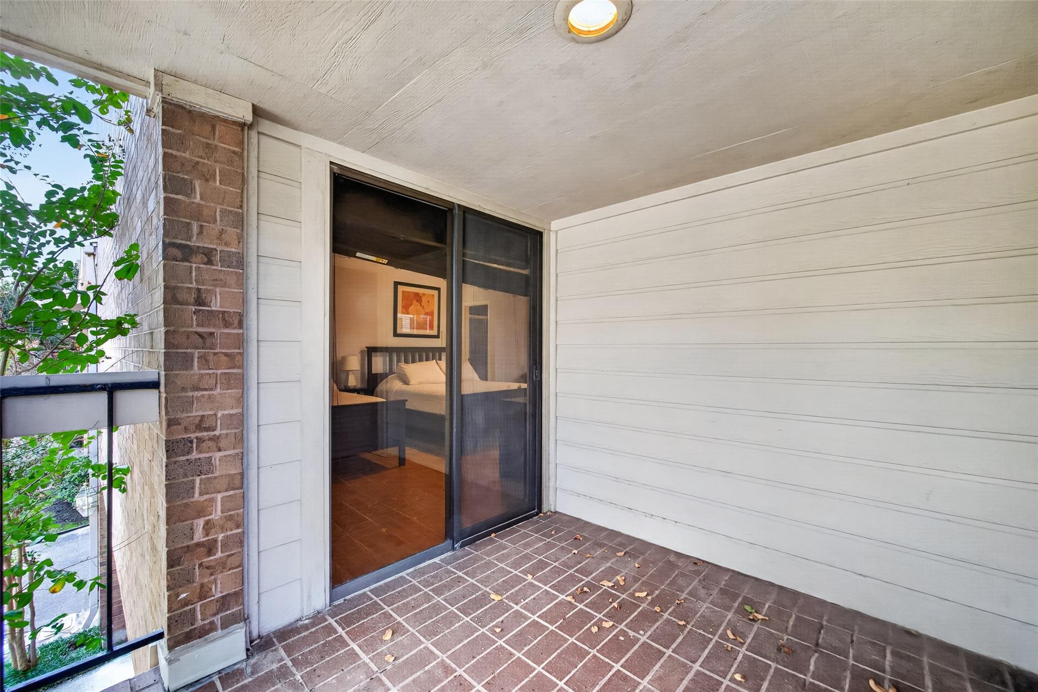 Covered balcony with brick-tile flooring and a sliding glass door looking into a furnished bedroom.