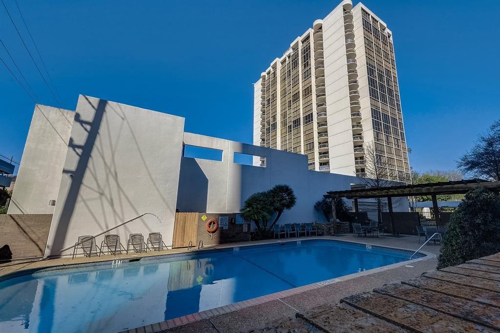 Outdoor swimming pool in front of a tall apartment tower, with lounge chairs and a shaded seating area.