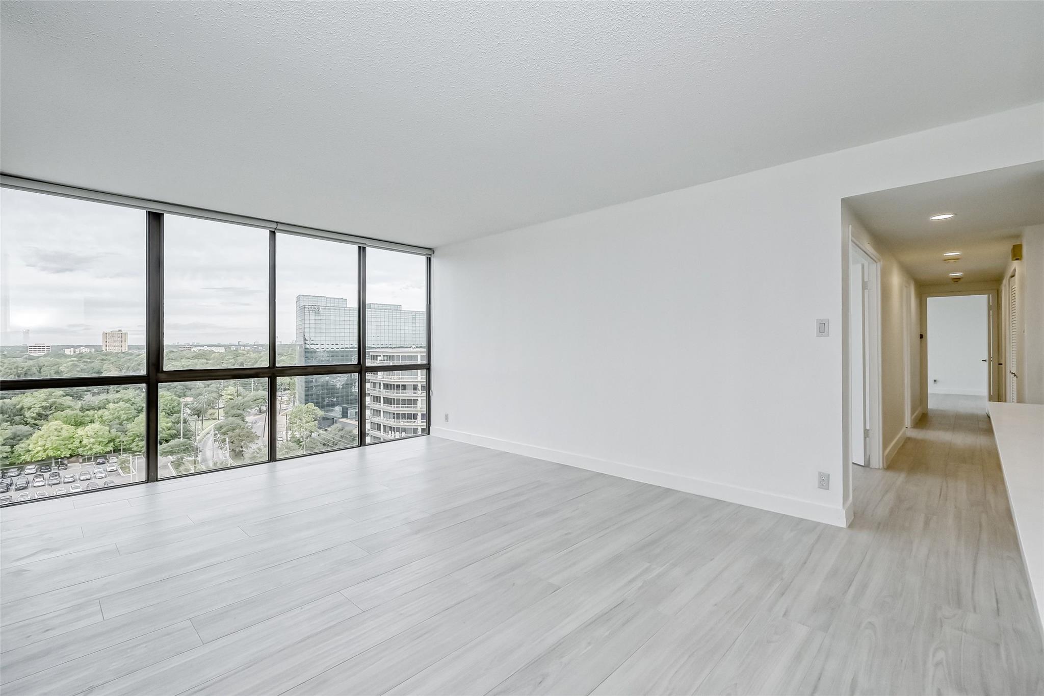 Empty condo living area with floor-to-ceiling windows overlooking trees and high-rise buildings.