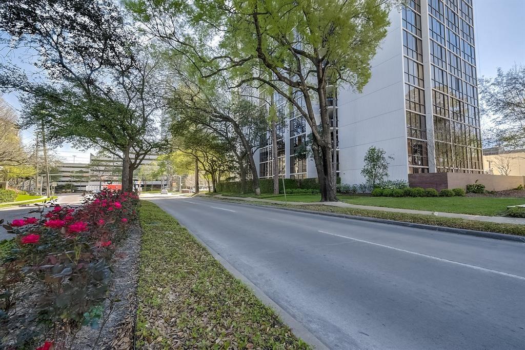 Multi-story residential building beside a tree-lined road and landscaped flower beds.