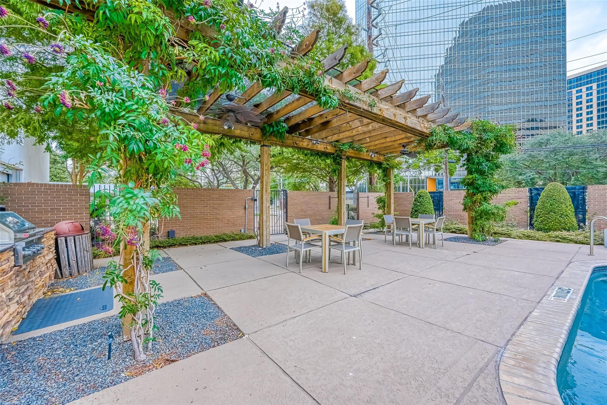 Poolside patio with a vine-covered pergola, outdoor dining tables, and city buildings beyond the brick wall.