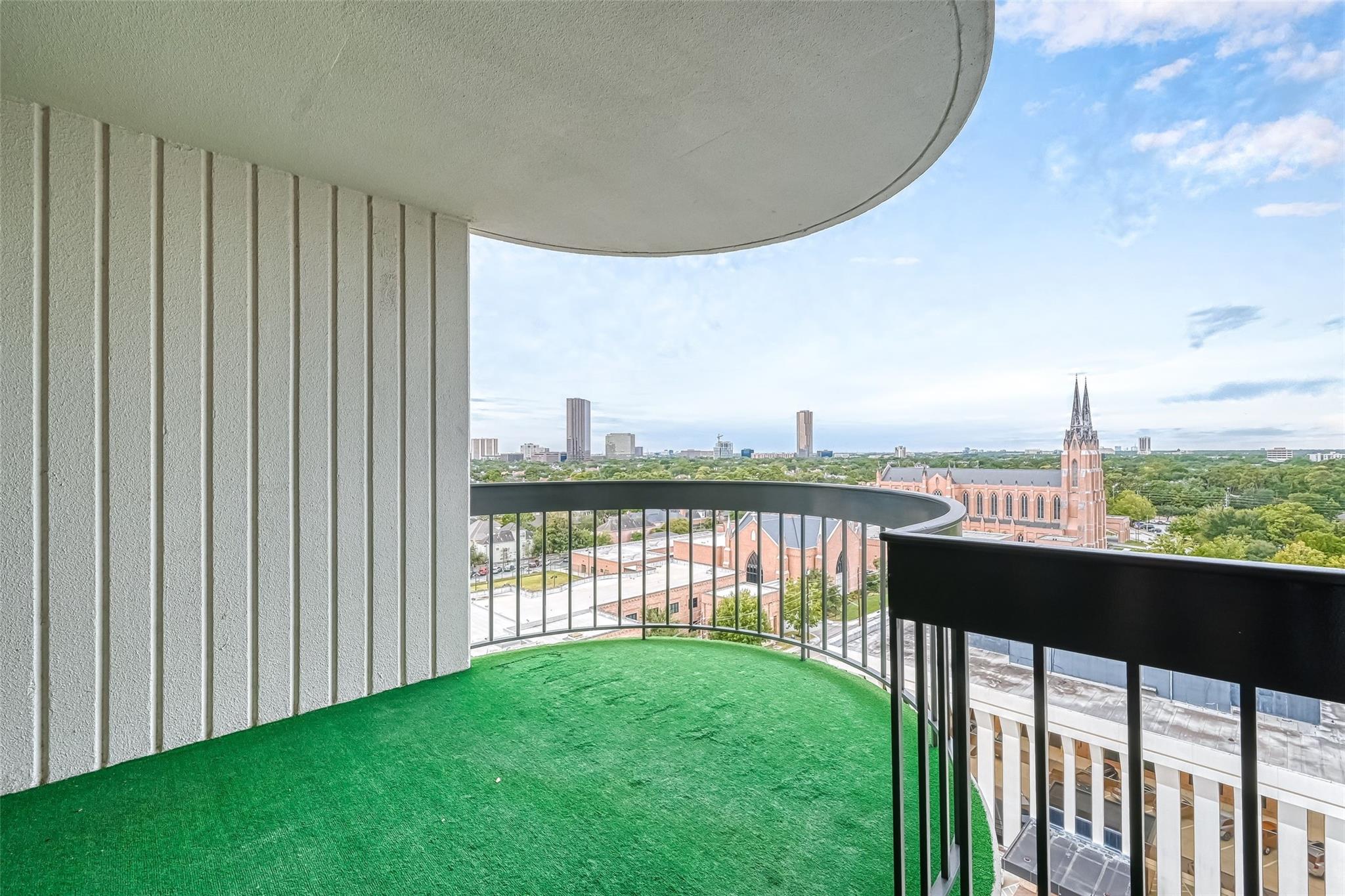 High-rise balcony with green turf and curved railings overlooking a city skyline and a red-brick church.