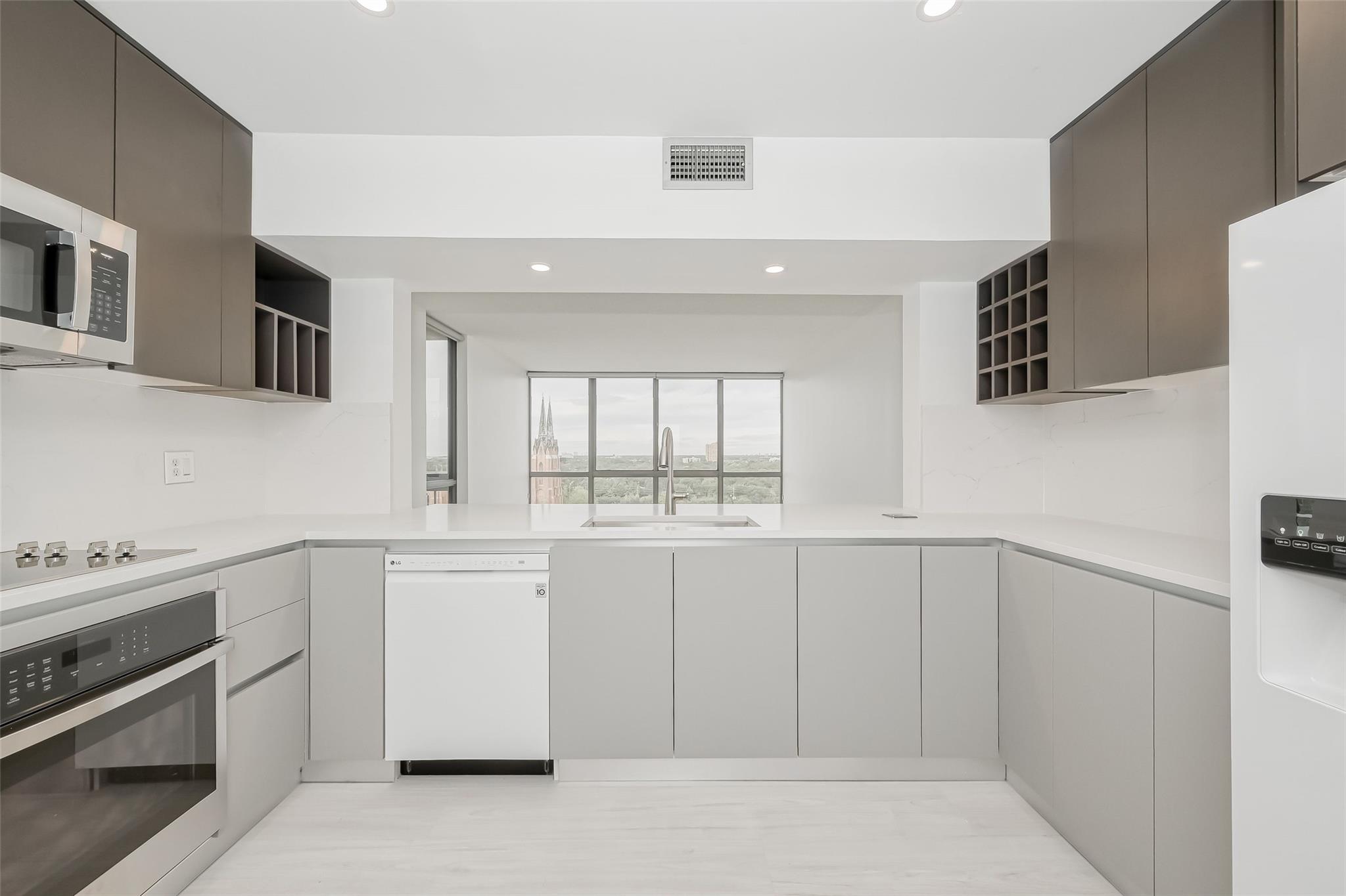 Modern kitchen with gray cabinets, white counters, and a window overlooking greenery and a church spire.