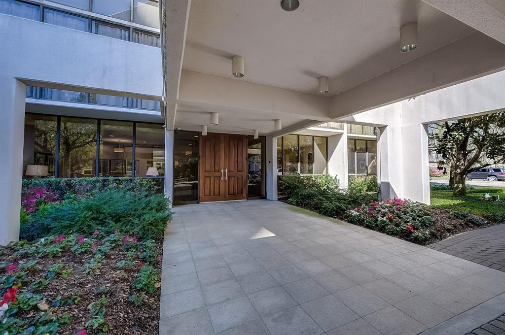 Residential building entrance with wood double doors, a covered tiled walkway, and flowering landscaping.