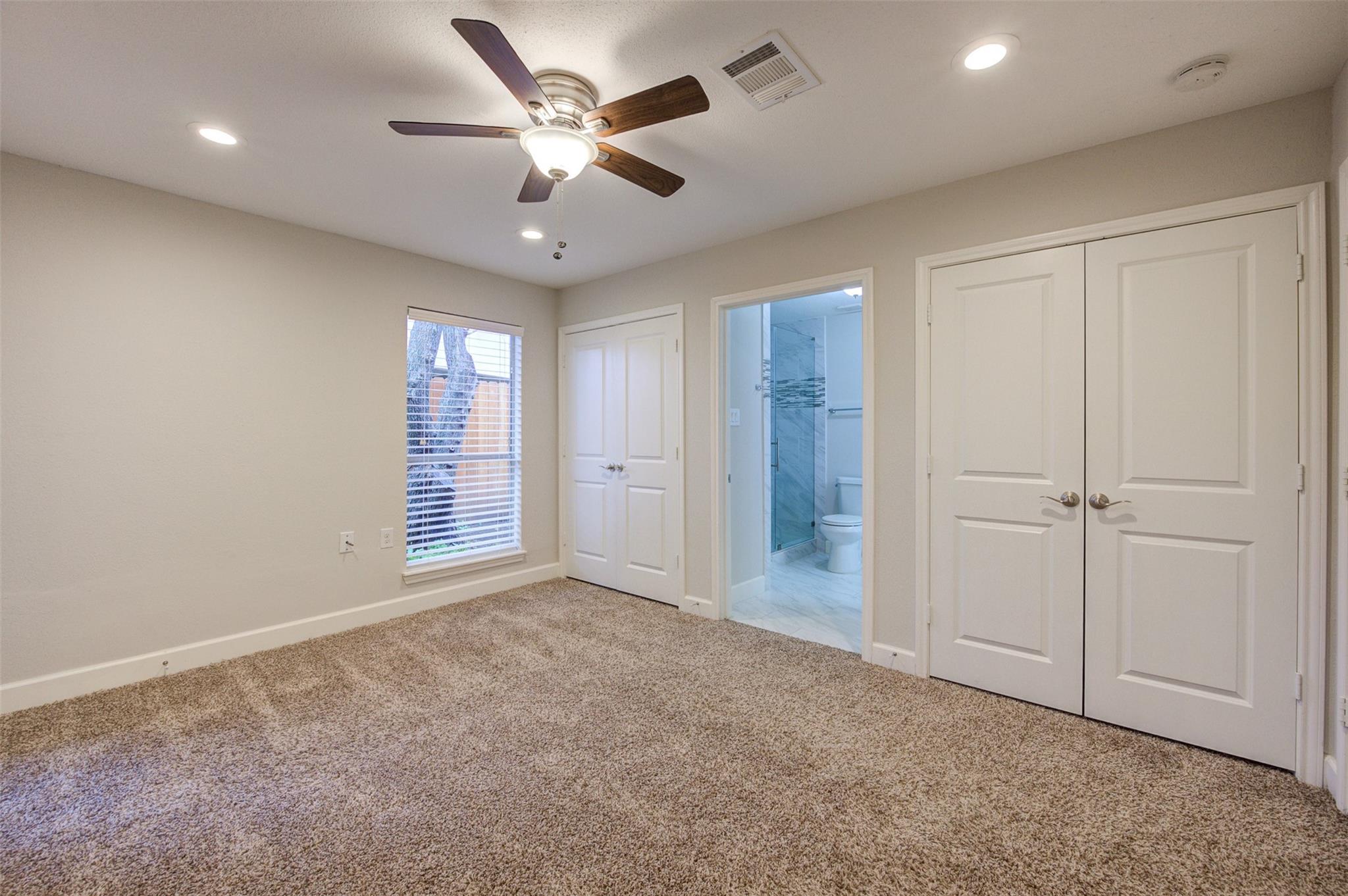 Empty carpeted bedroom with a ceiling fan, white closet doors, and an adjoining bathroom.