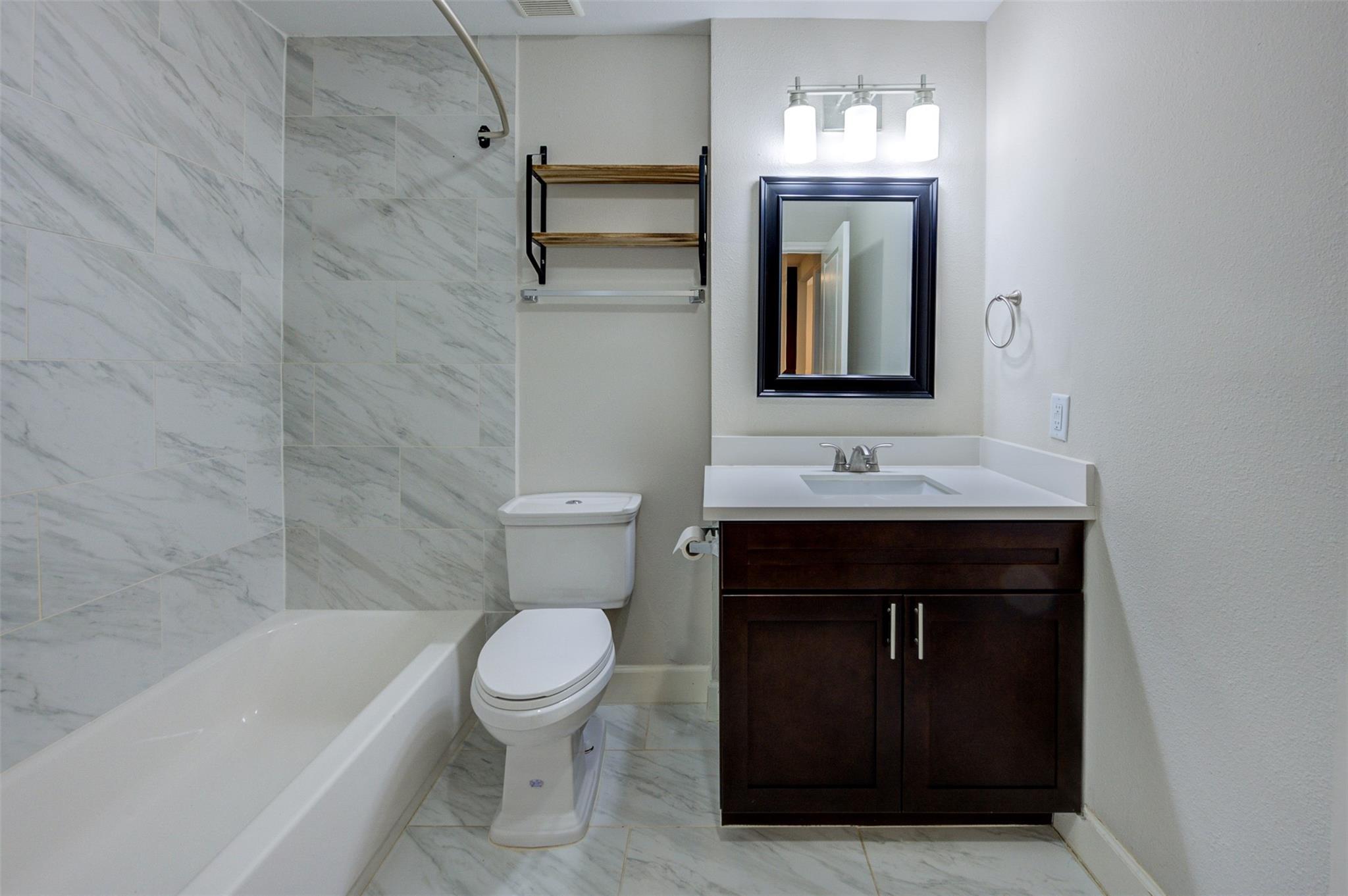 Bathroom with marble-look tile, bathtub, toilet, and dark wood vanity with a white sink.