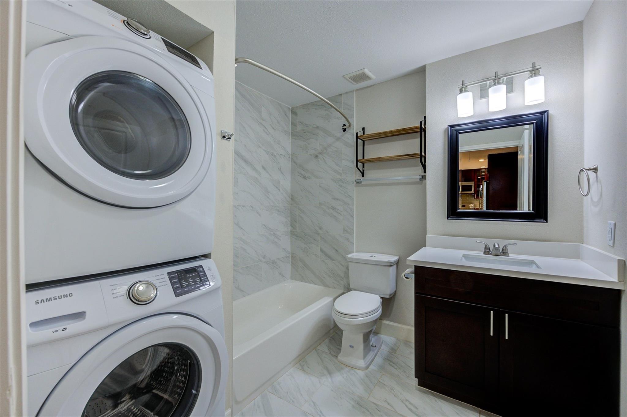 Bathroom with stacked washer and dryer, marble-look tub surround, and dark wood vanity.