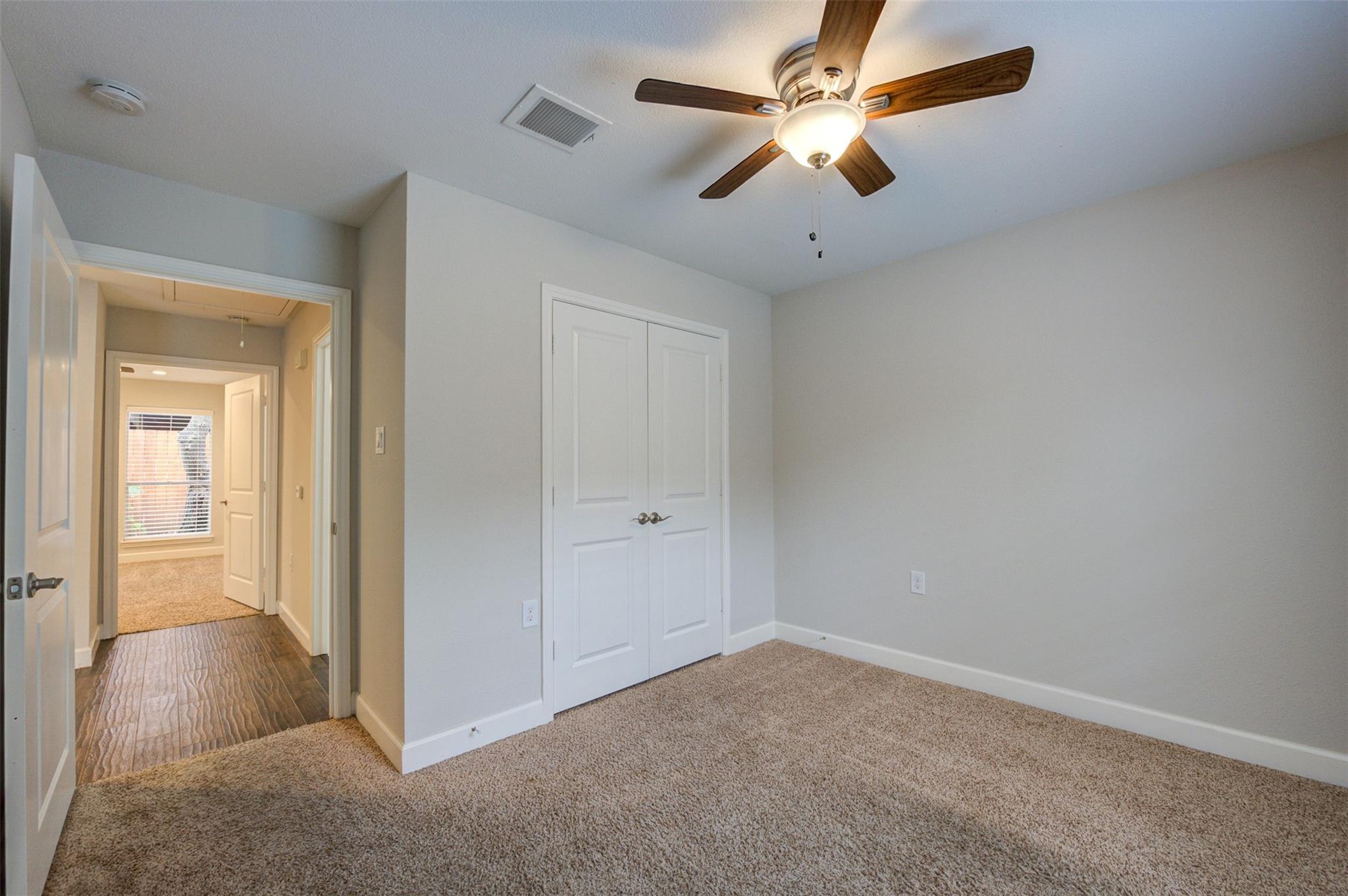 Empty carpeted bedroom with white double closet doors and a lit ceiling fan; doorway opens to a windowed room.