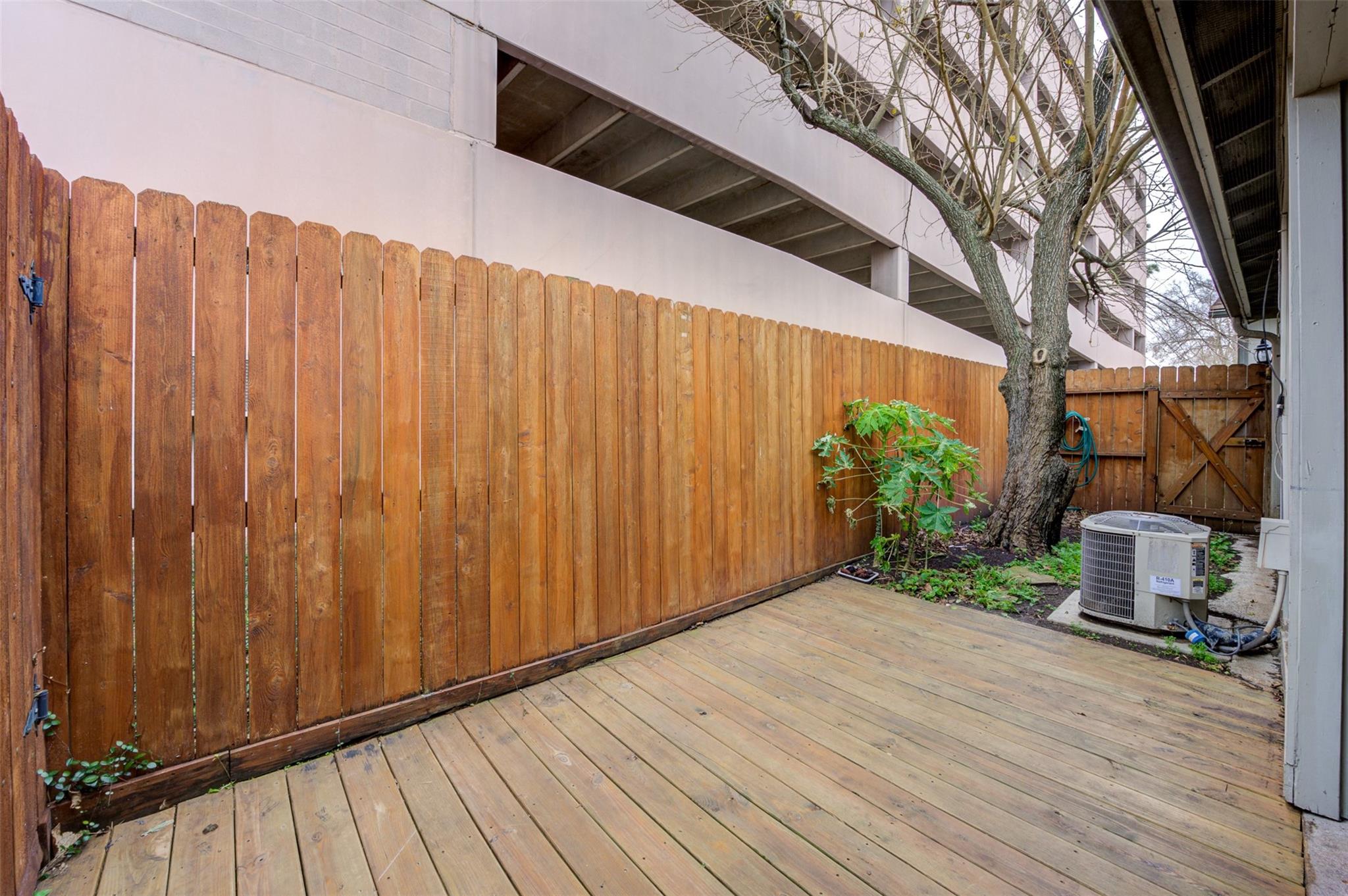 Private wooden deck patio with a tall privacy fence, a leafless tree, and green plants.