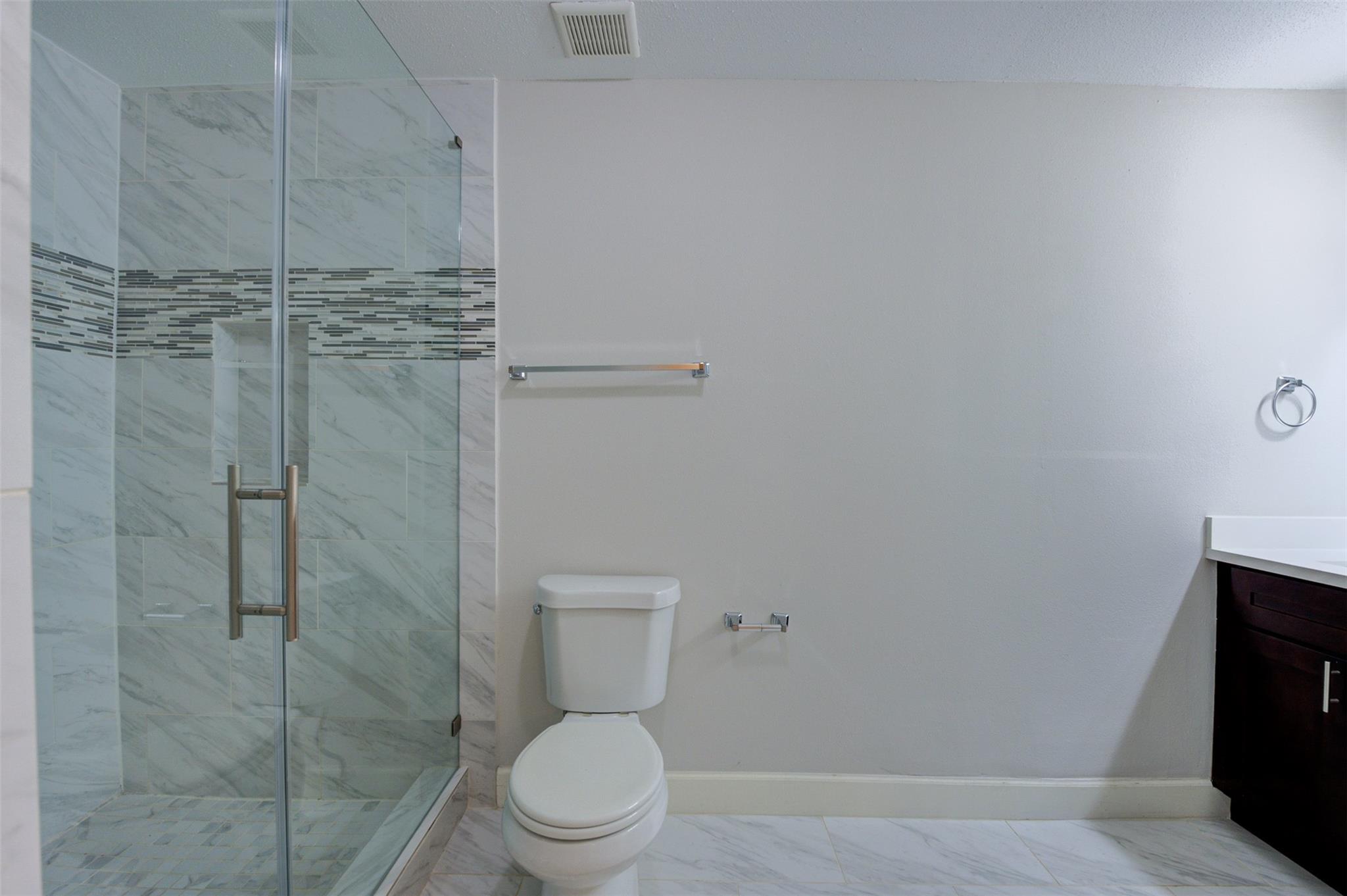 Bathroom with a glass shower, marble-look tile, mosaic accent band, toilet, and dark wood vanity.