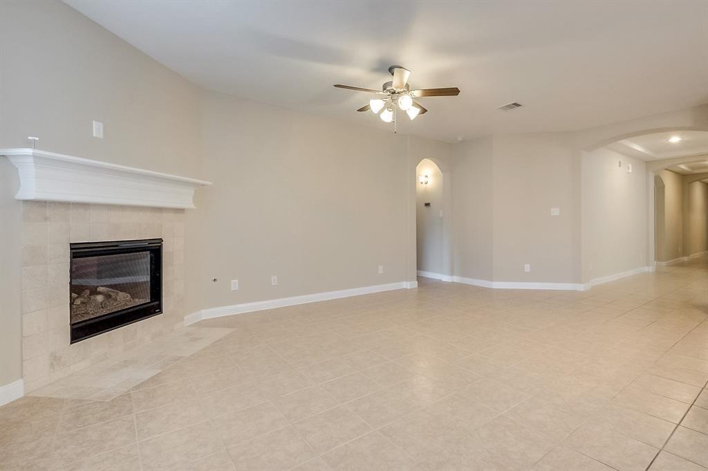 Empty living room with a tiled fireplace, beige tile floors, and a ceiling fan with lights.