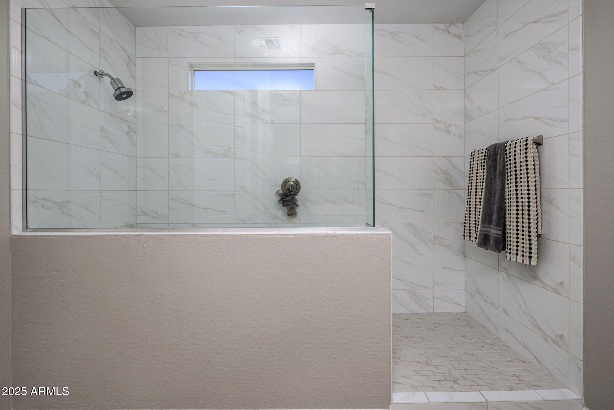 Walk-in shower with marble-look tile, glass partition, narrow window, and patterned towels.