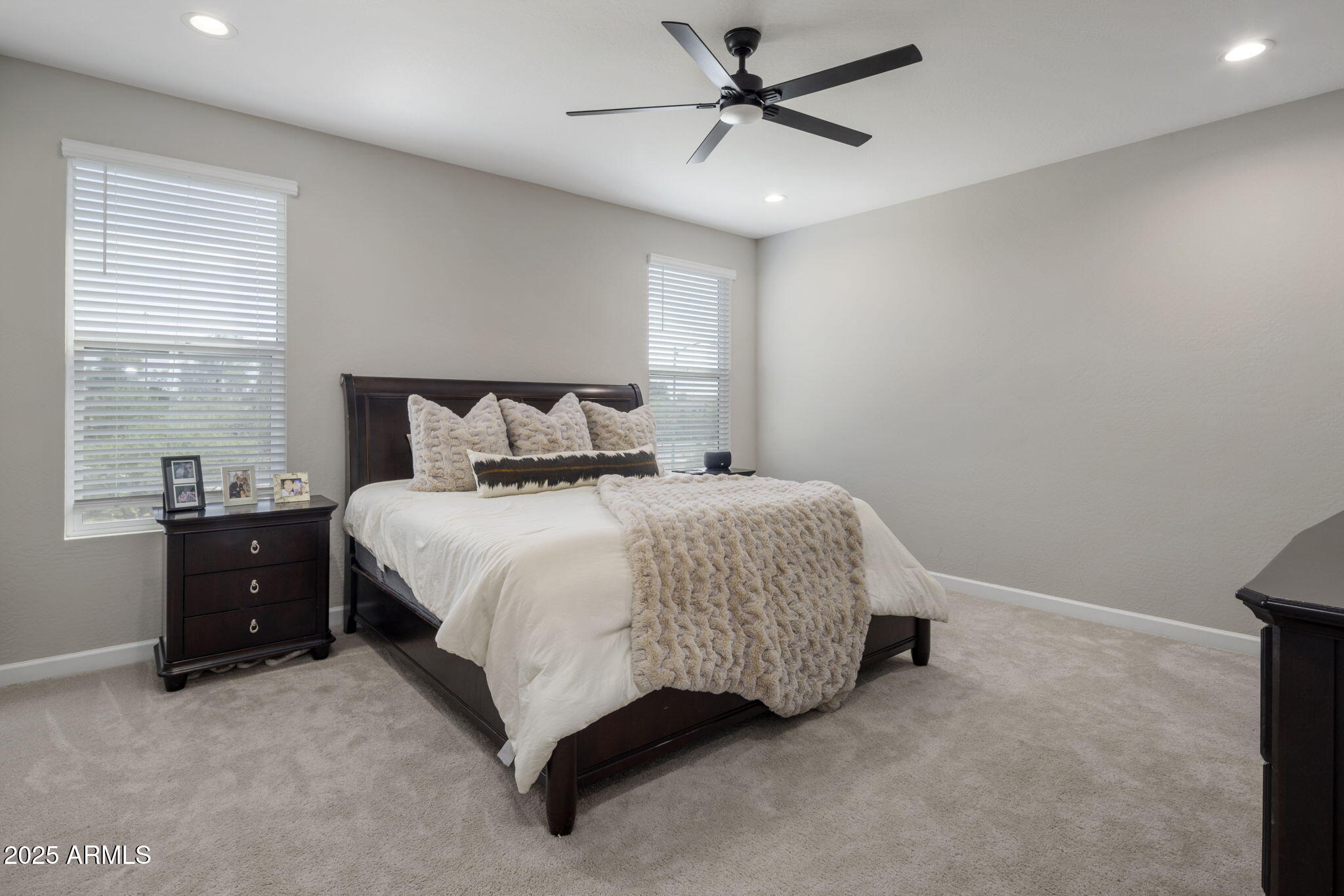 Furnished bedroom with a dark wood bed, cream textured bedding, beige carpet, and two windows with blinds.