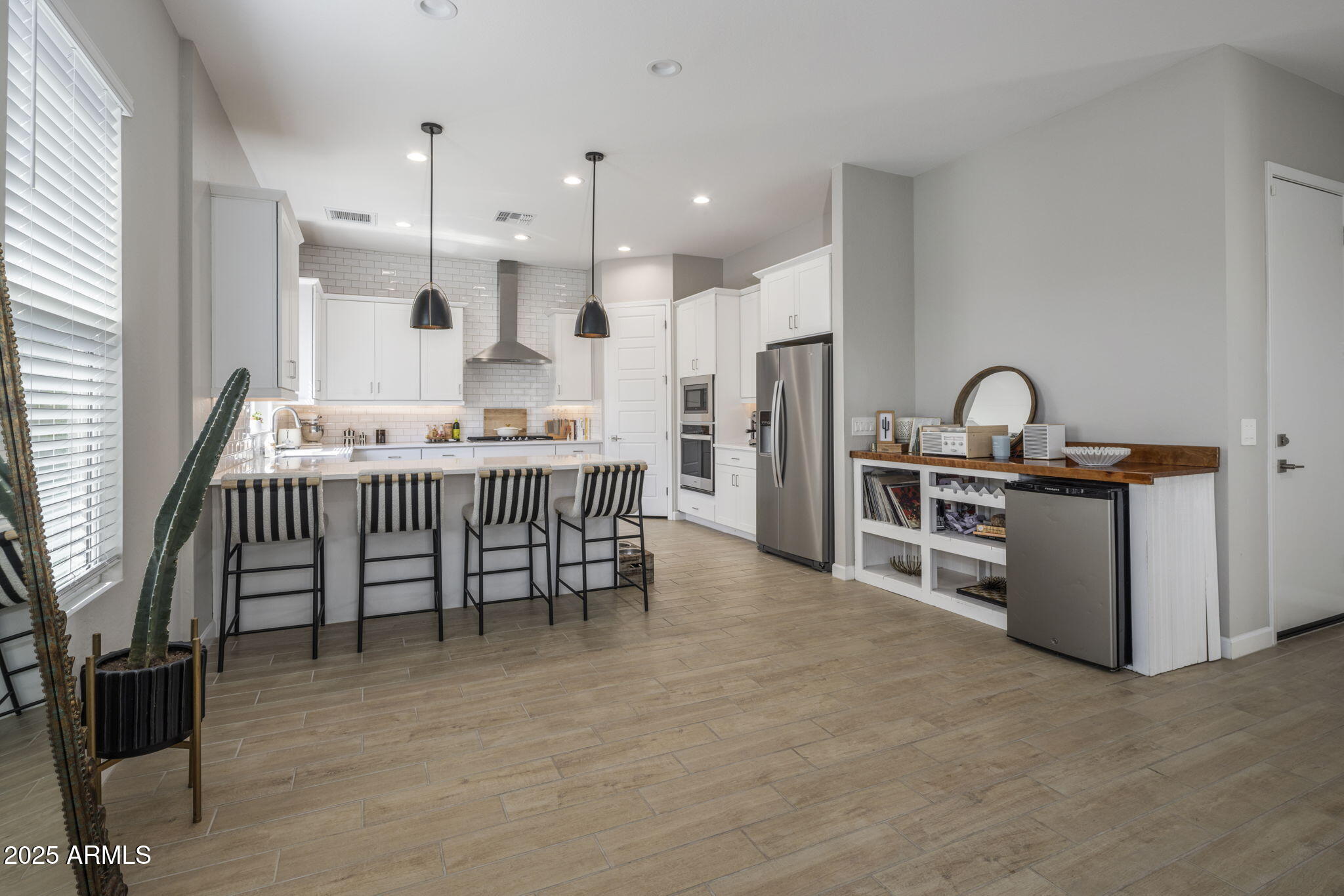Modern white kitchen with a large island, striped bar stools, stainless appliances, and a tall cactus by the window.