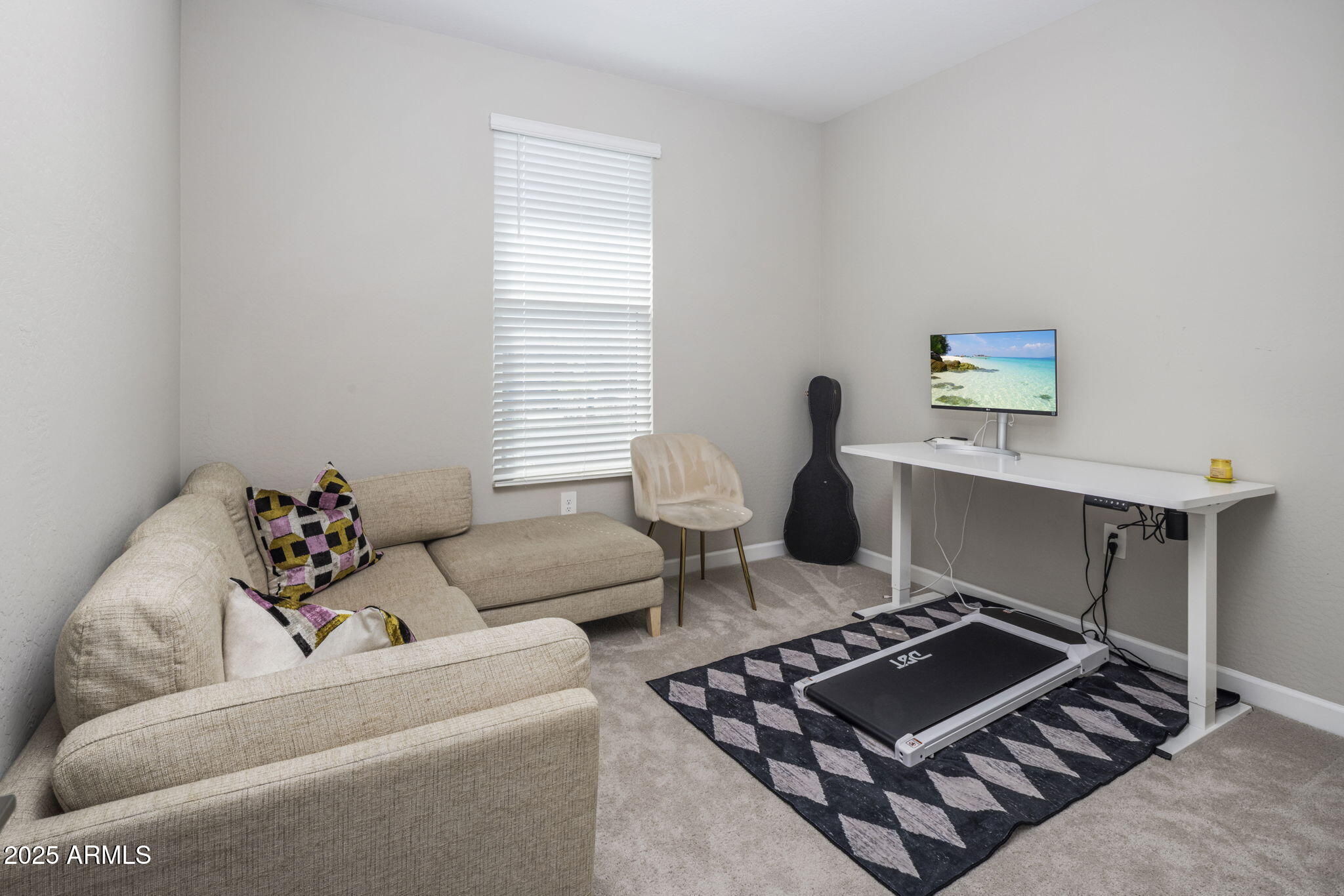 Home office with a beige sectional, standing desk, treadmill, and guitar case.