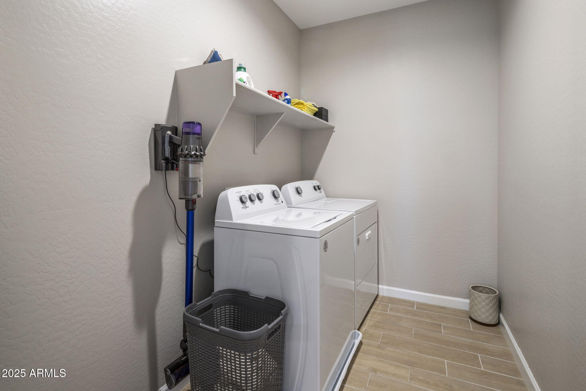 Laundry room with a washer and dryer, wall-mounted vacuum, and shelf of cleaning supplies.