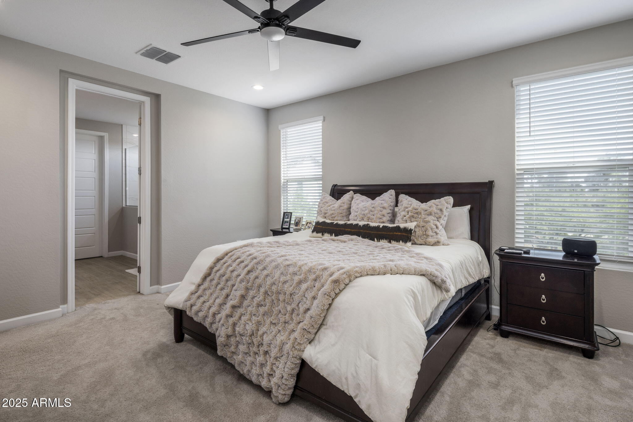 Bedroom with a dark wood bed, textured beige bedding, ceiling fan, and two windows with blinds.