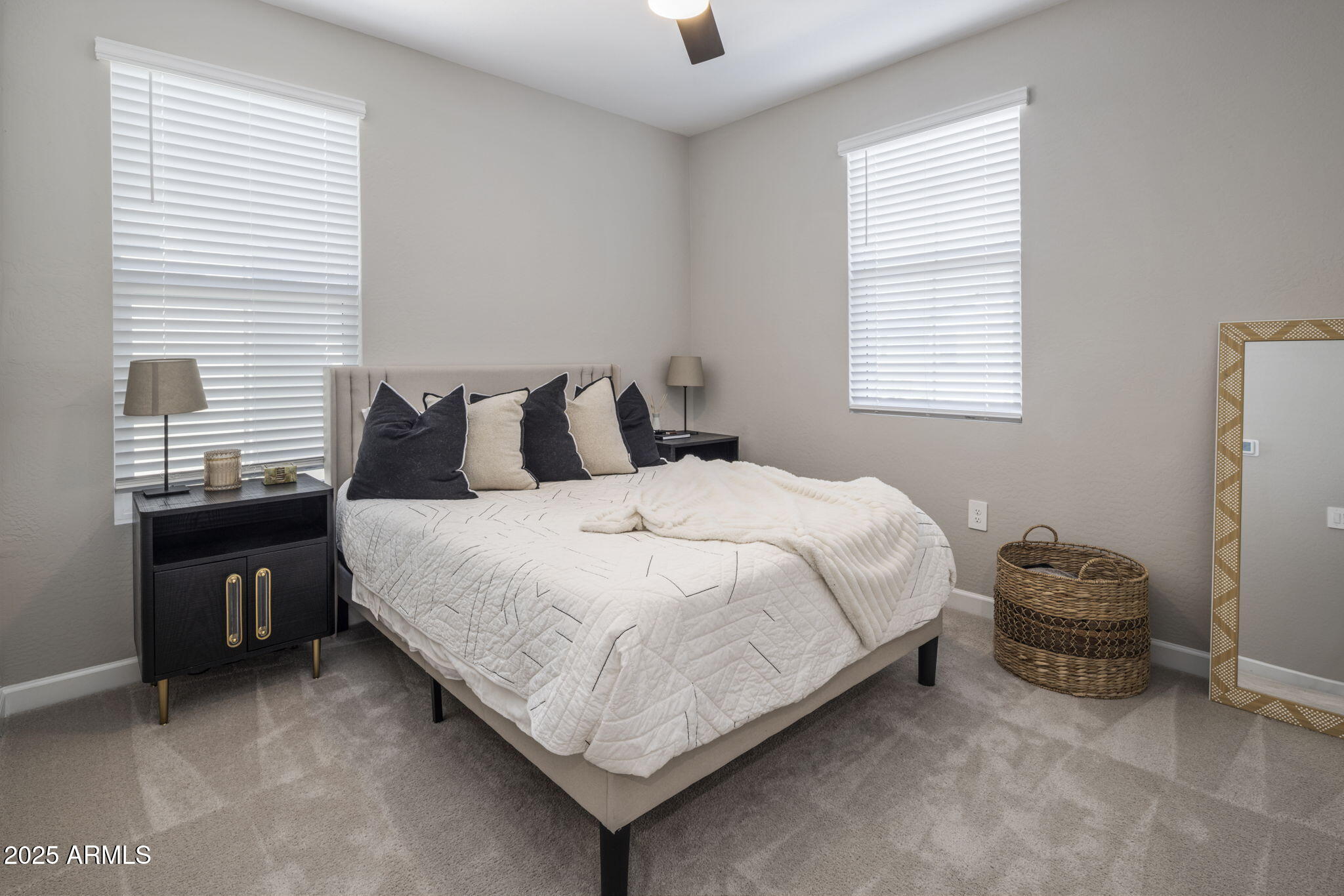 Furnished bedroom with a white-quilted bed, dark bedside cabinet, and two windows with white blinds.