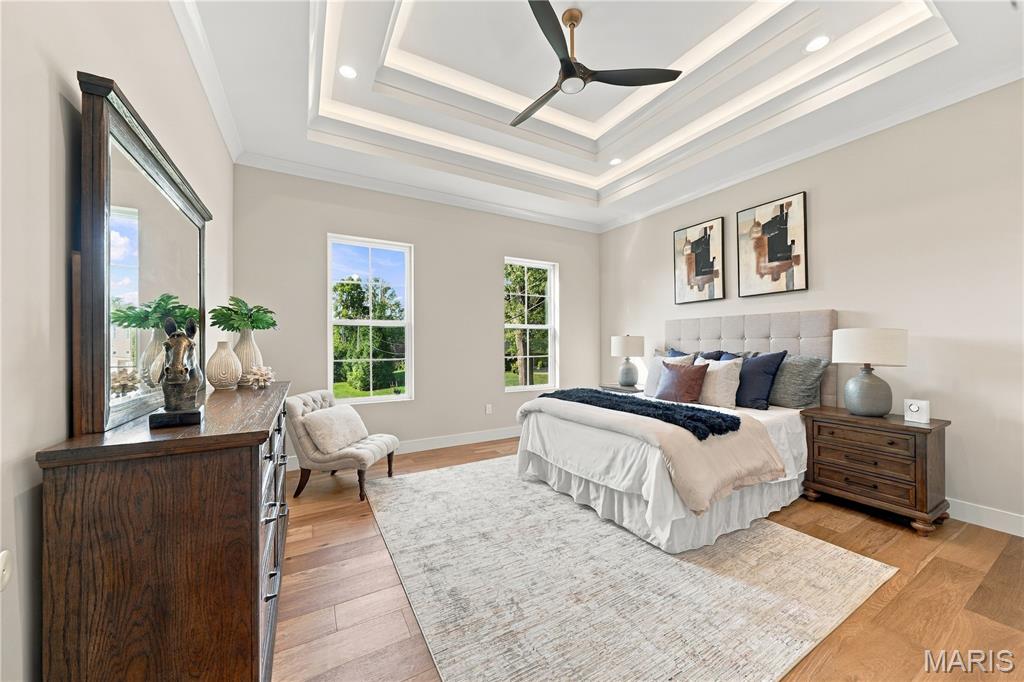Bedroom with a large bed beneath a tray ceiling, hardwood floors, and tall windows overlooking trees.