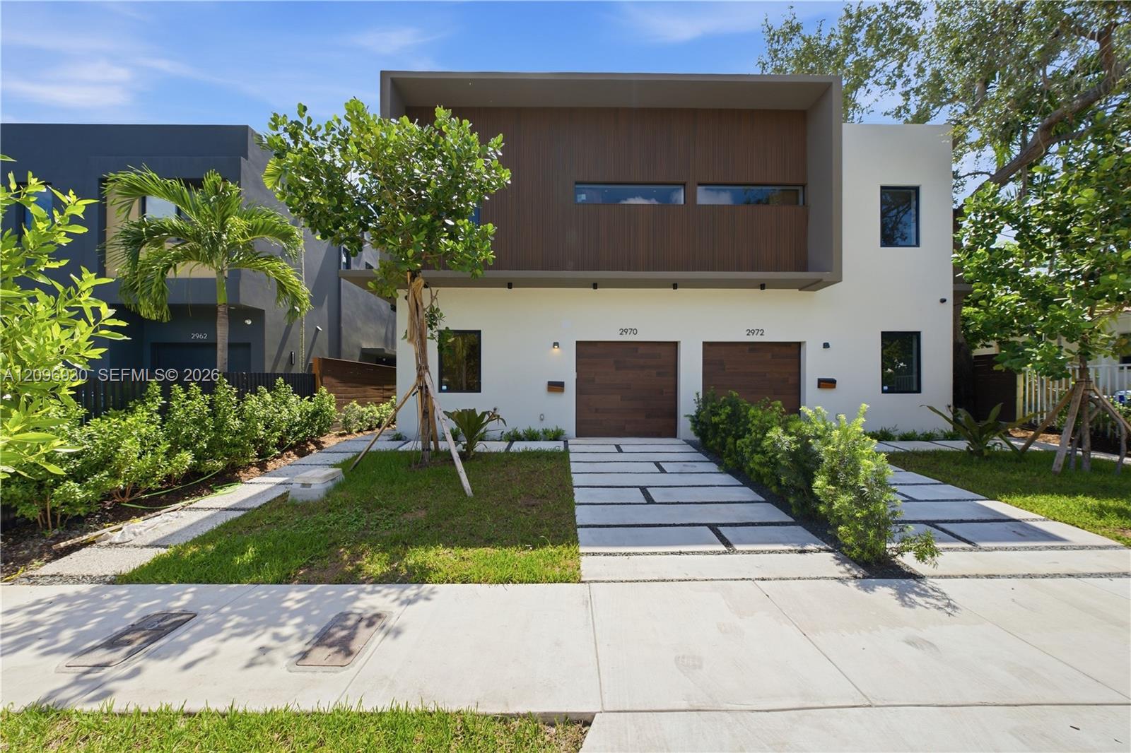 Modern white residence with brown upper facade, two wood-toned garage doors, and a landscaped paver driveway.
