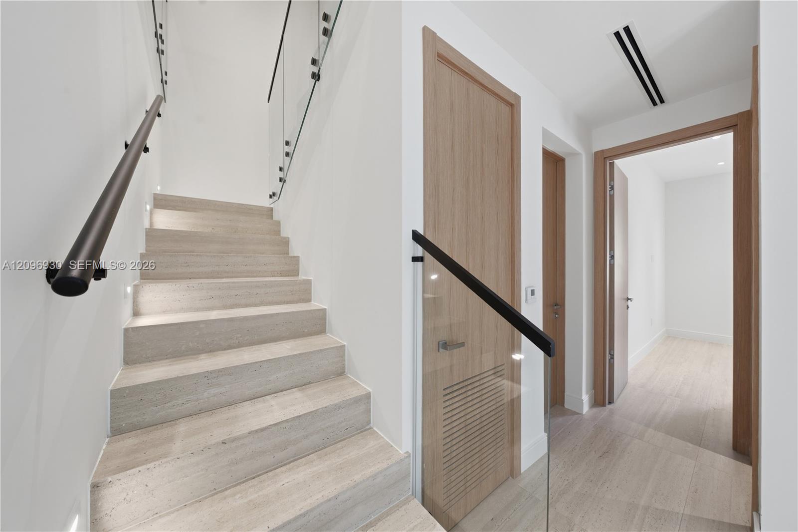 Pale stone staircase with glass railing and black handrail beside a light wood-trimmed hallway.