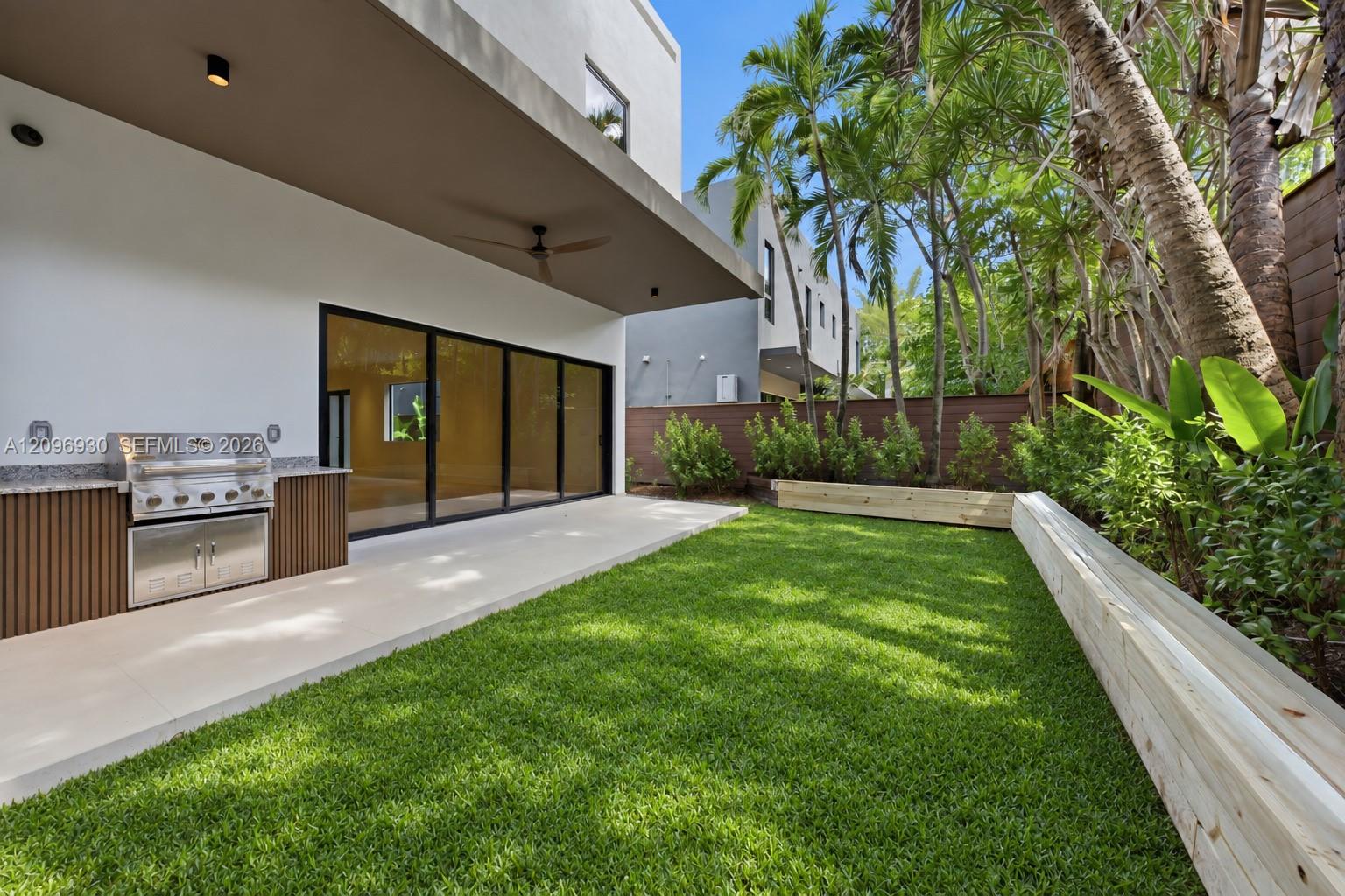 Modern home patio with an outdoor kitchen, grassy lawn, raised beds, and tropical palms.
