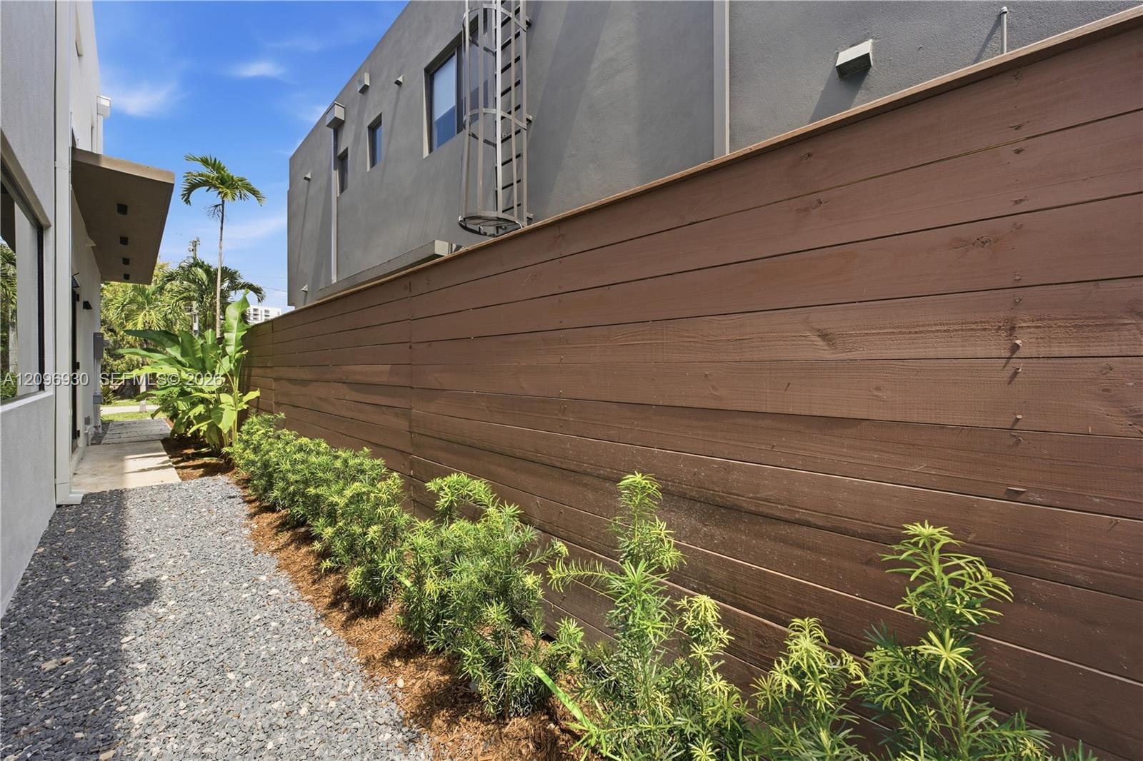 Modern home side yard with a long wooden fence, gravel path, and tropical plants.
