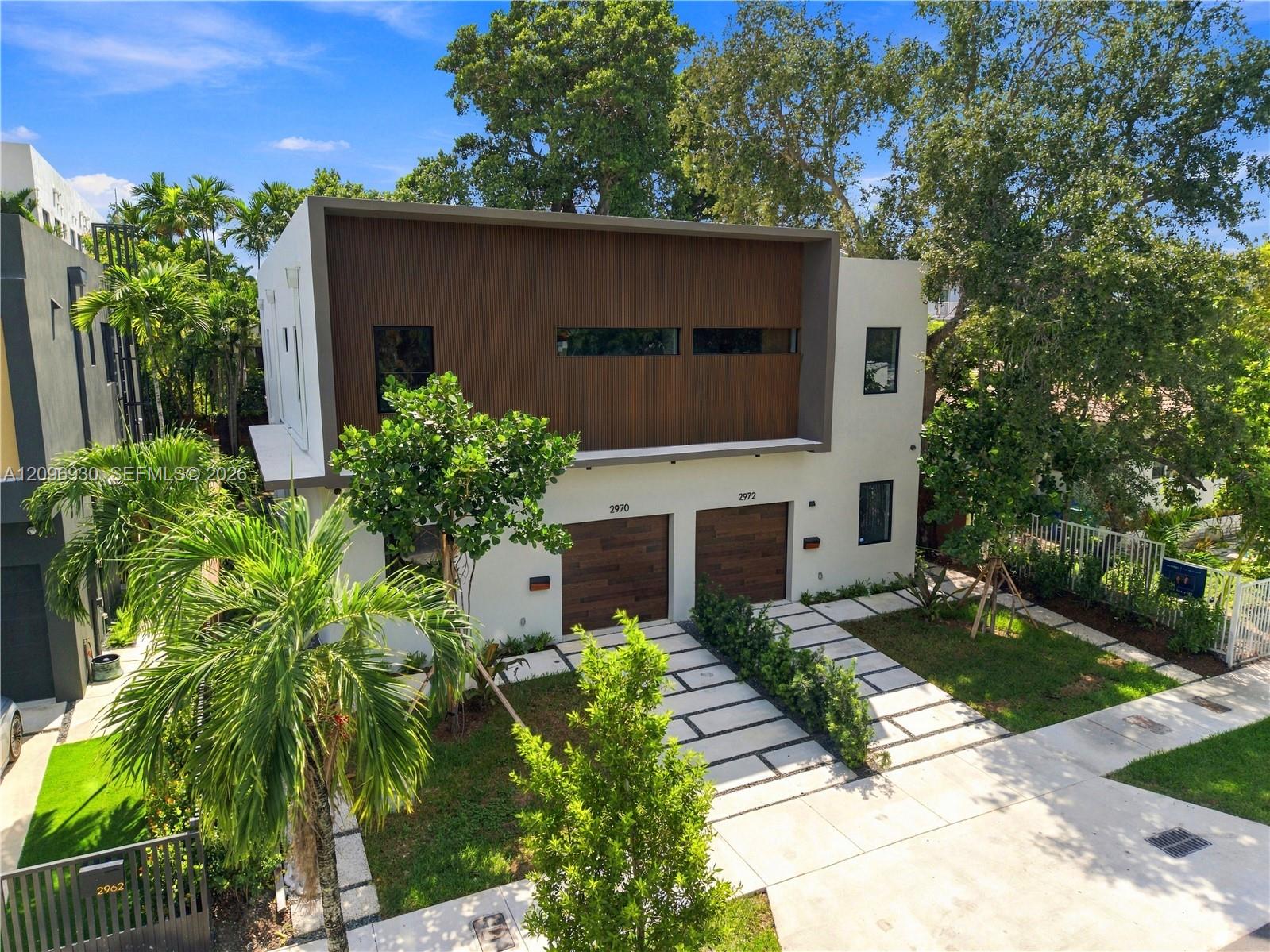 Modern white residence with a wood-clad upper facade, paired garage doors, and tropical landscaping.