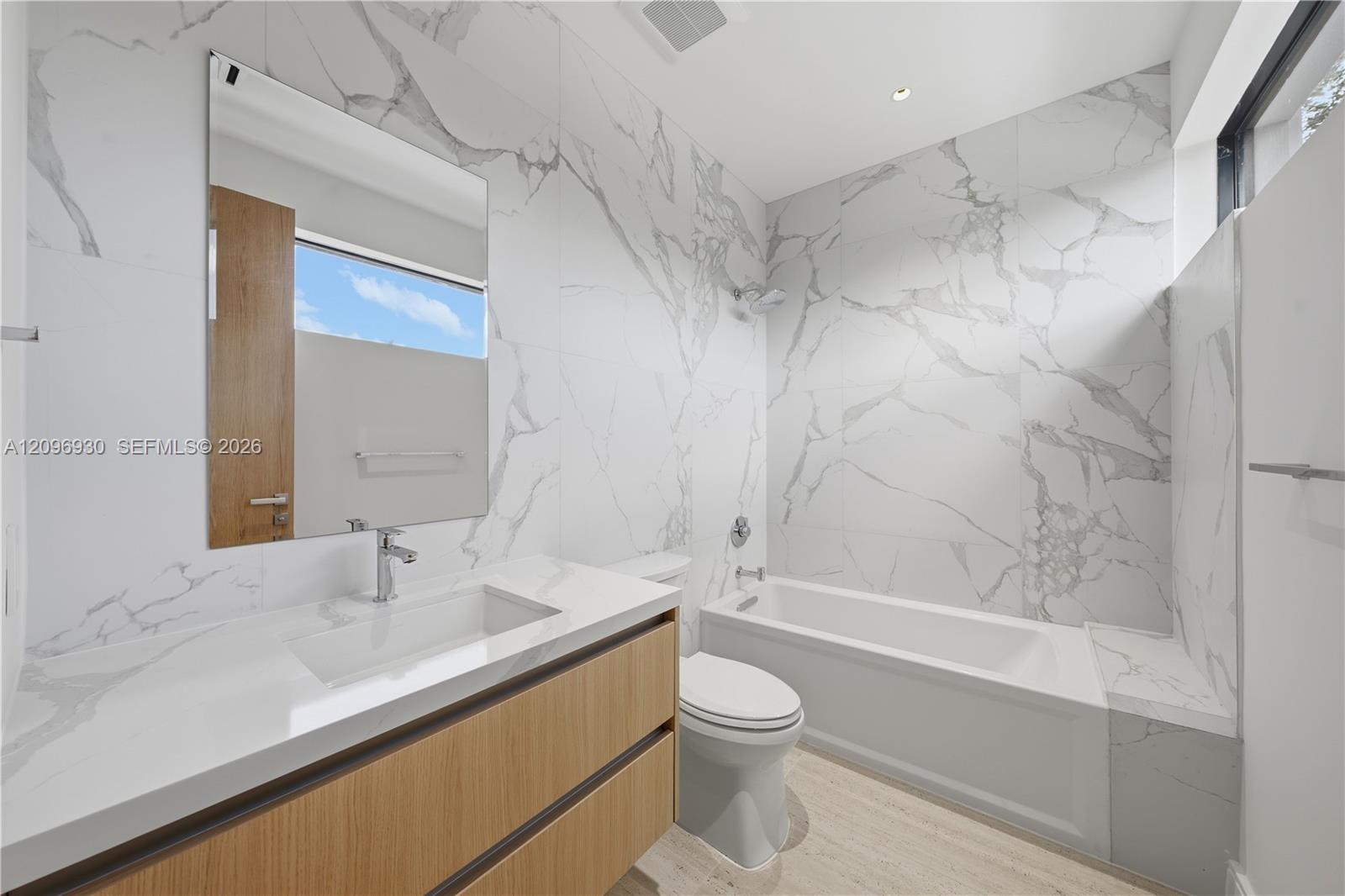 Modern bathroom with marble-look tile, a wood-front vanity, and a white bathtub.