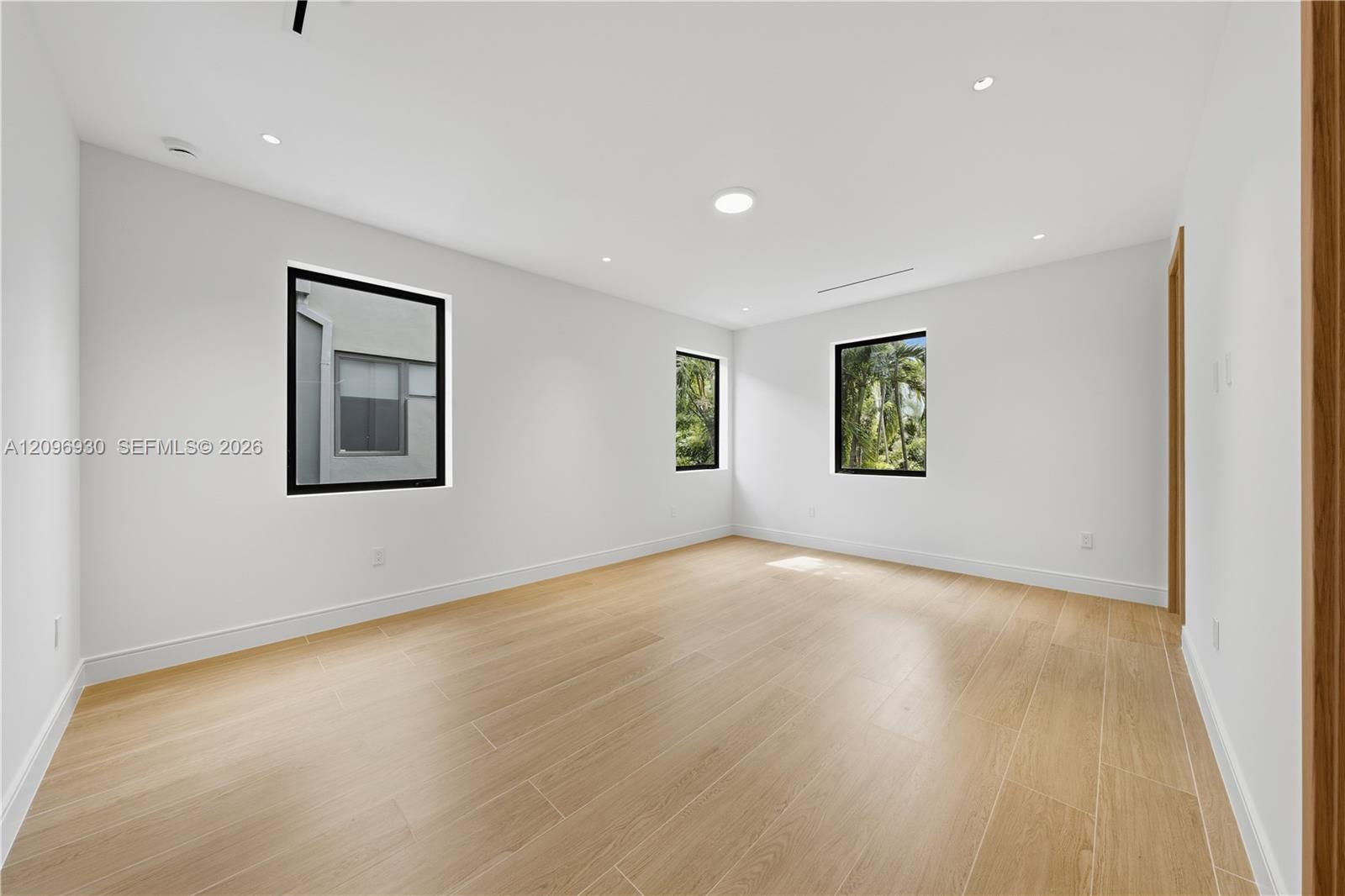 Empty room with light wood-look tile floors, recessed lights, and windows overlooking greenery.