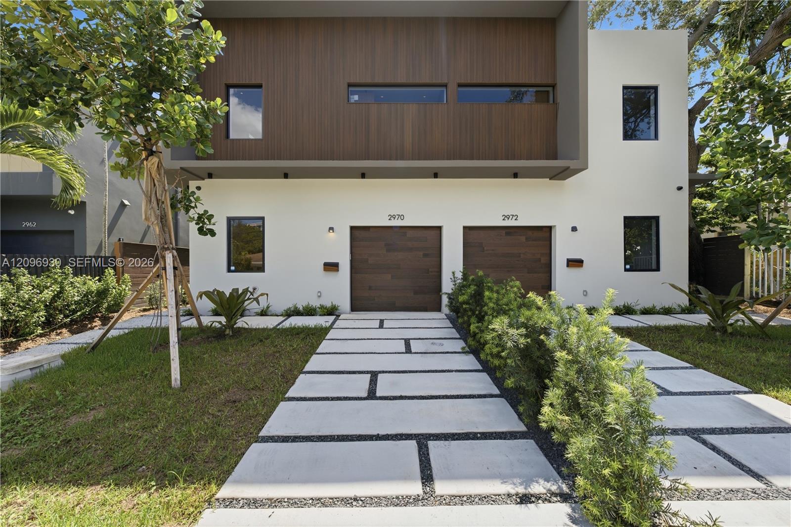 Modern white two-story residence with dark vertical siding, paired wood-look garage doors, and a paver walkway.