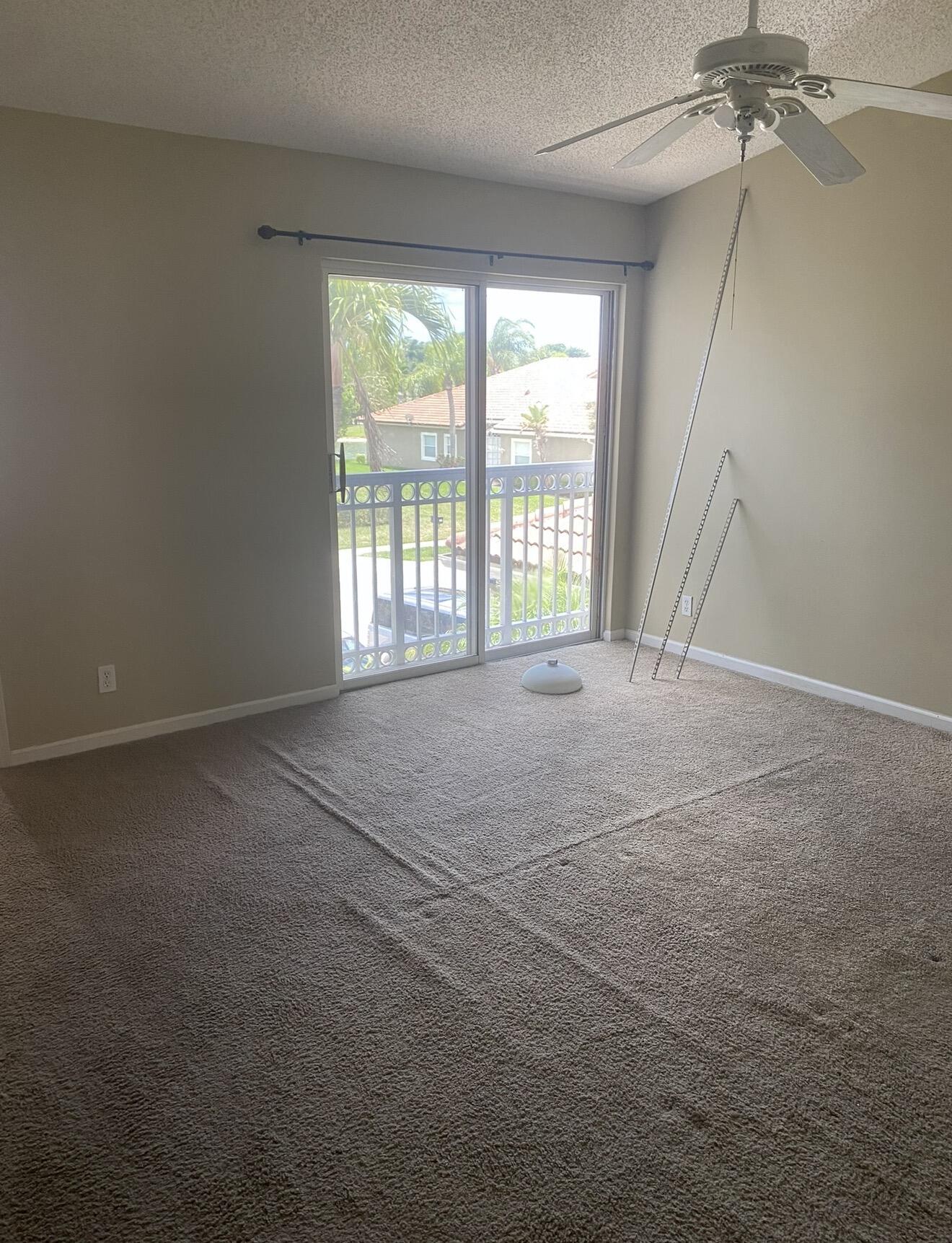 Empty carpeted room with sliding balcony doors, a ceiling fan, and palm trees outside.
