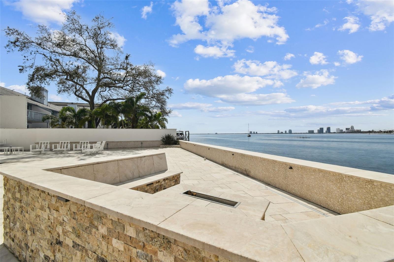 Waterfront terrace with pale stone paving, outdoor chairs, and a bay and city skyline view.