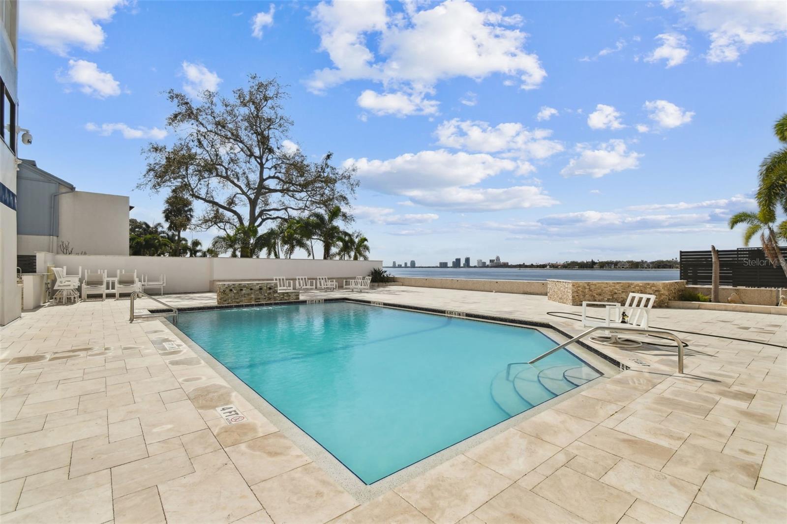 Outdoor pool with stone deck and seating overlooks the bay and distant city skyline.