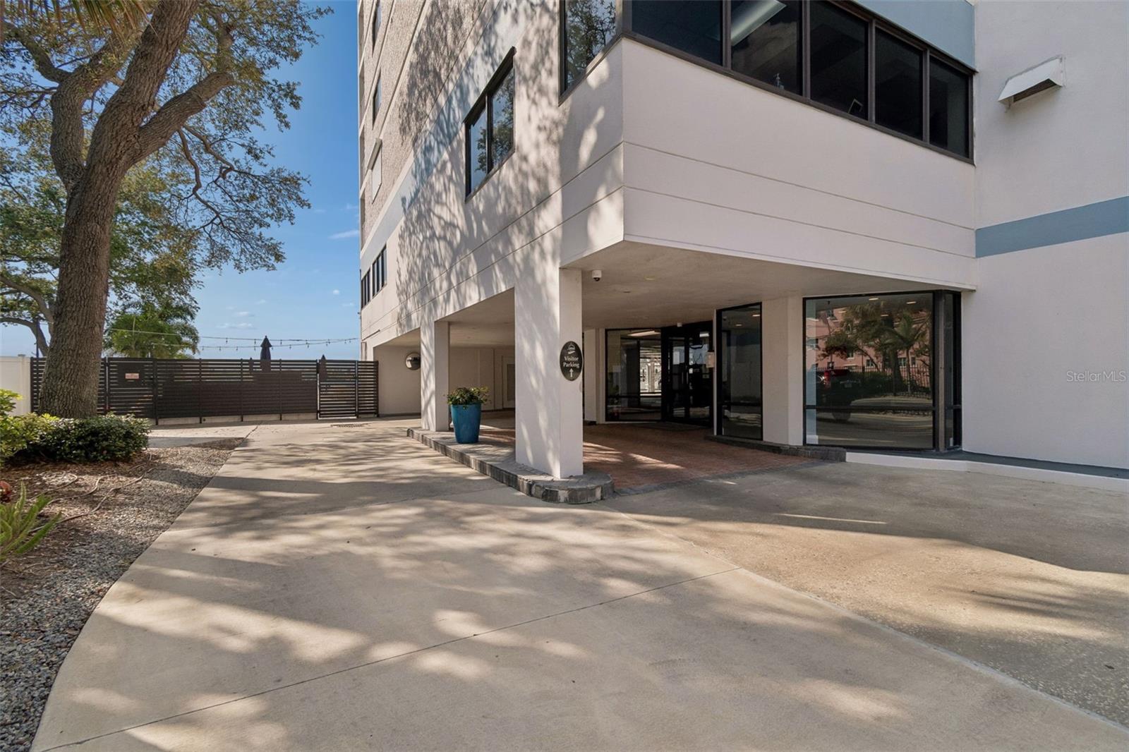 Condominium entrance with a covered drop-off area and gated driveway beside a large tree.